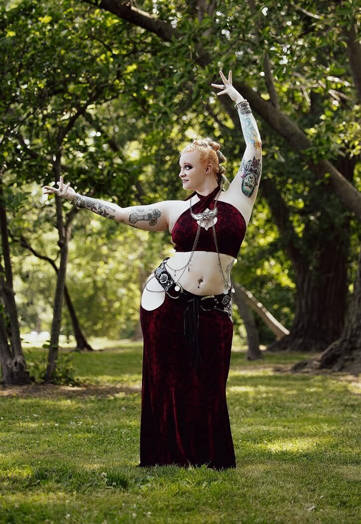 Person with tattoos in a long velvet dress dancing outdoors with arms raised, surrounded by trees and greenery.