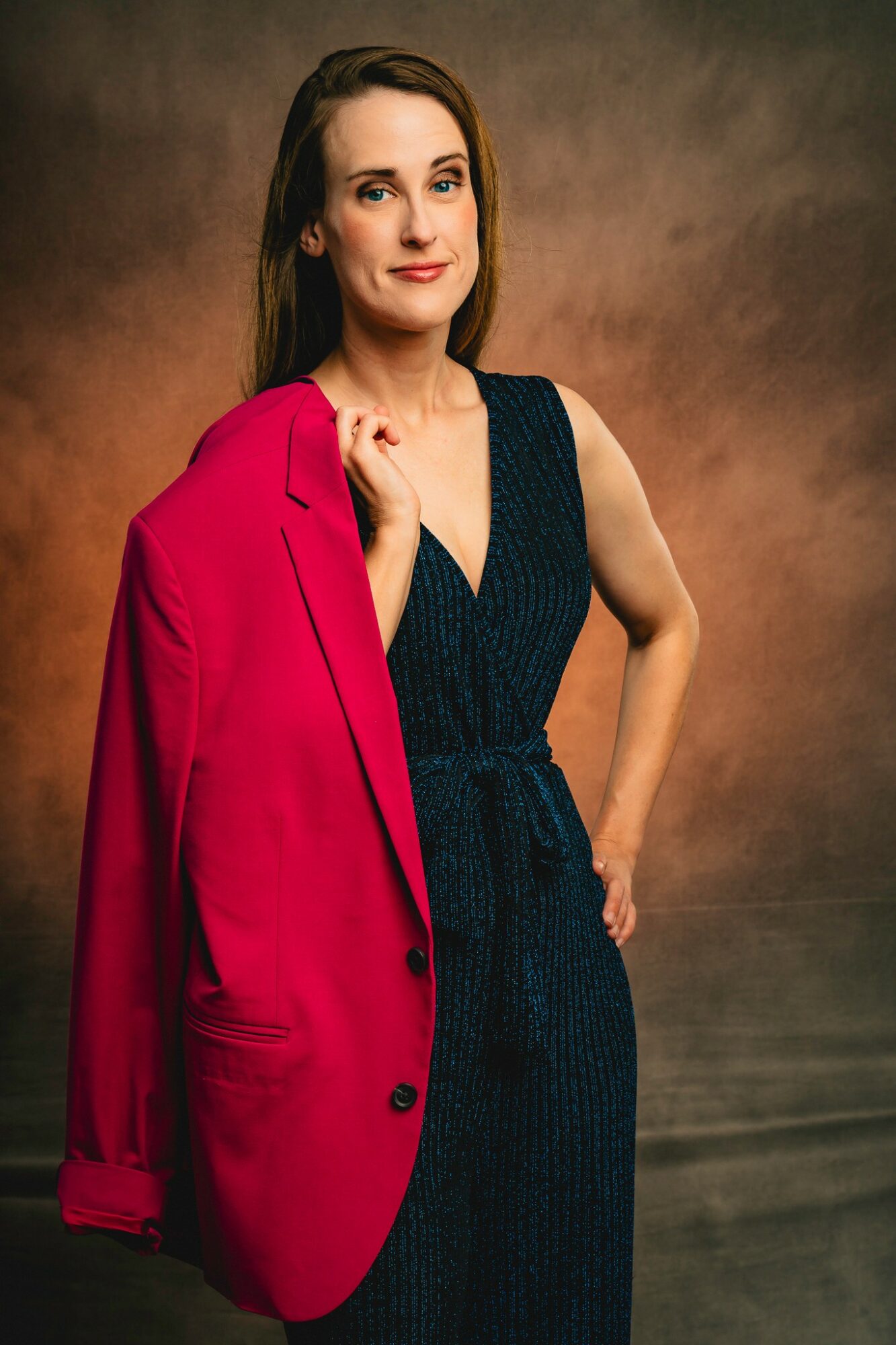 Woman with long brown hair wearing a dark blue dress and a red blazer over her shoulder, standing against a warm background.