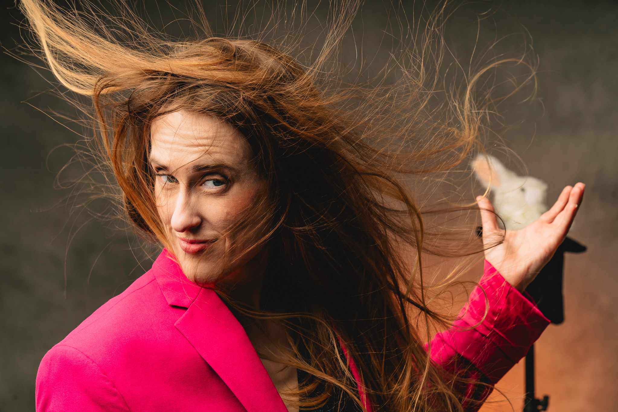 Woman with long hair holding a small white mouse, hair blowing, wearing a pink jacket, against a dark background.