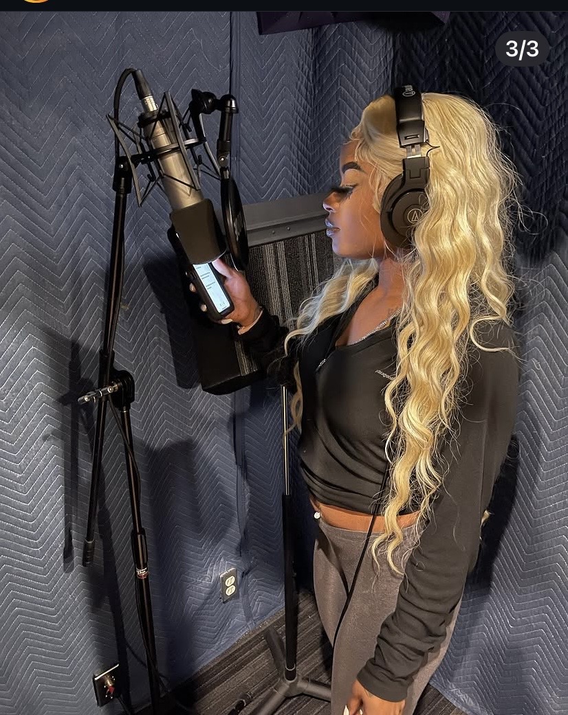Woman with long blonde curly hair wearing headphones, standing in a recording studio, reading from a tablet in front of a microphone.