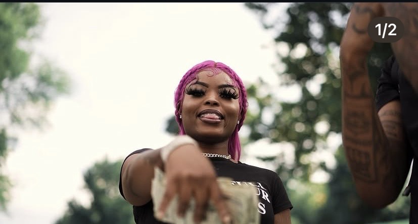Woman with pink hair smiling outdoors, pointing towards the camera, with trees in the background.