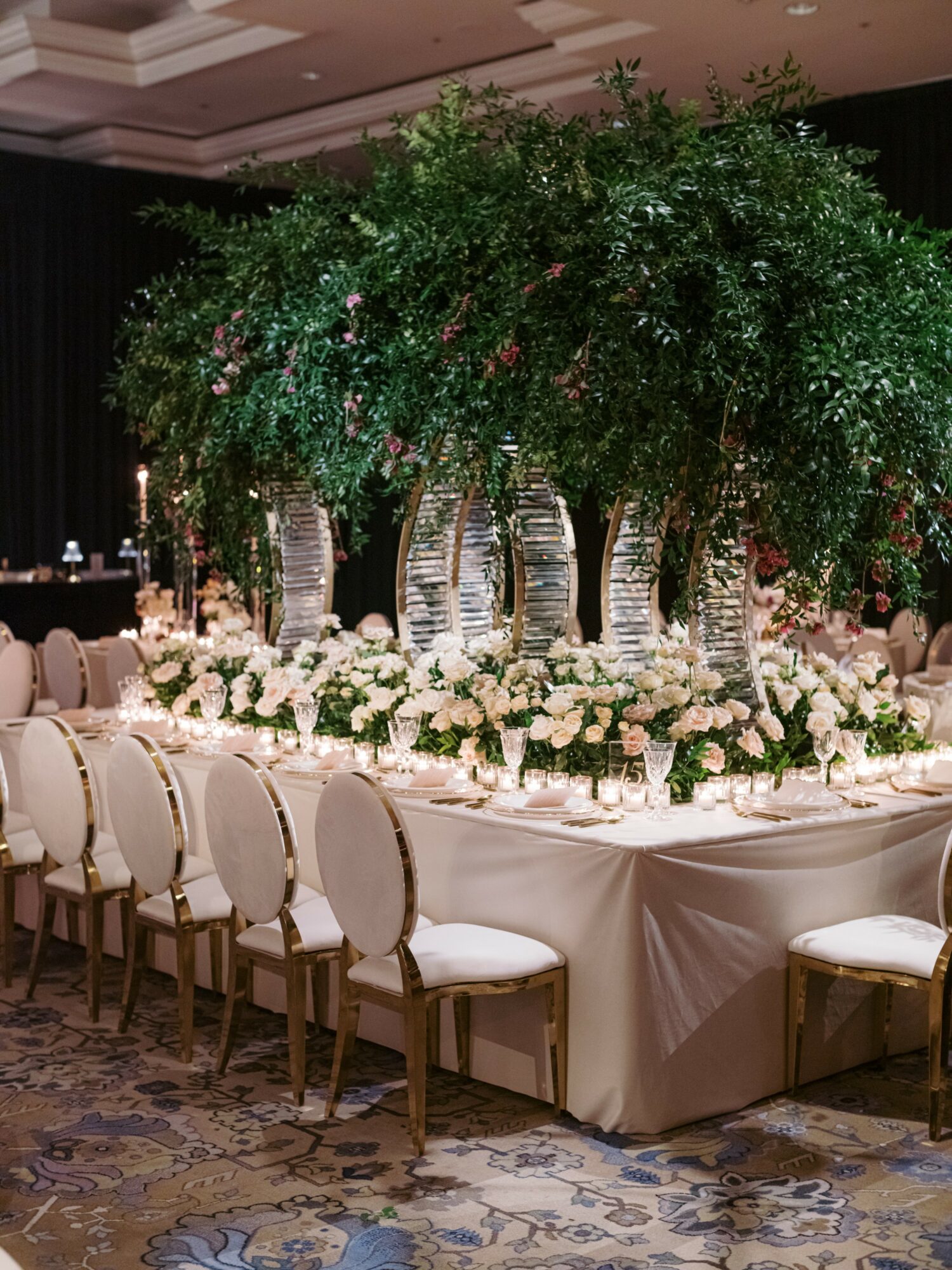 Elegant banquet table with a large floral centerpiece of trees and white flowers, surrounded by chairs in a decorated event hall.