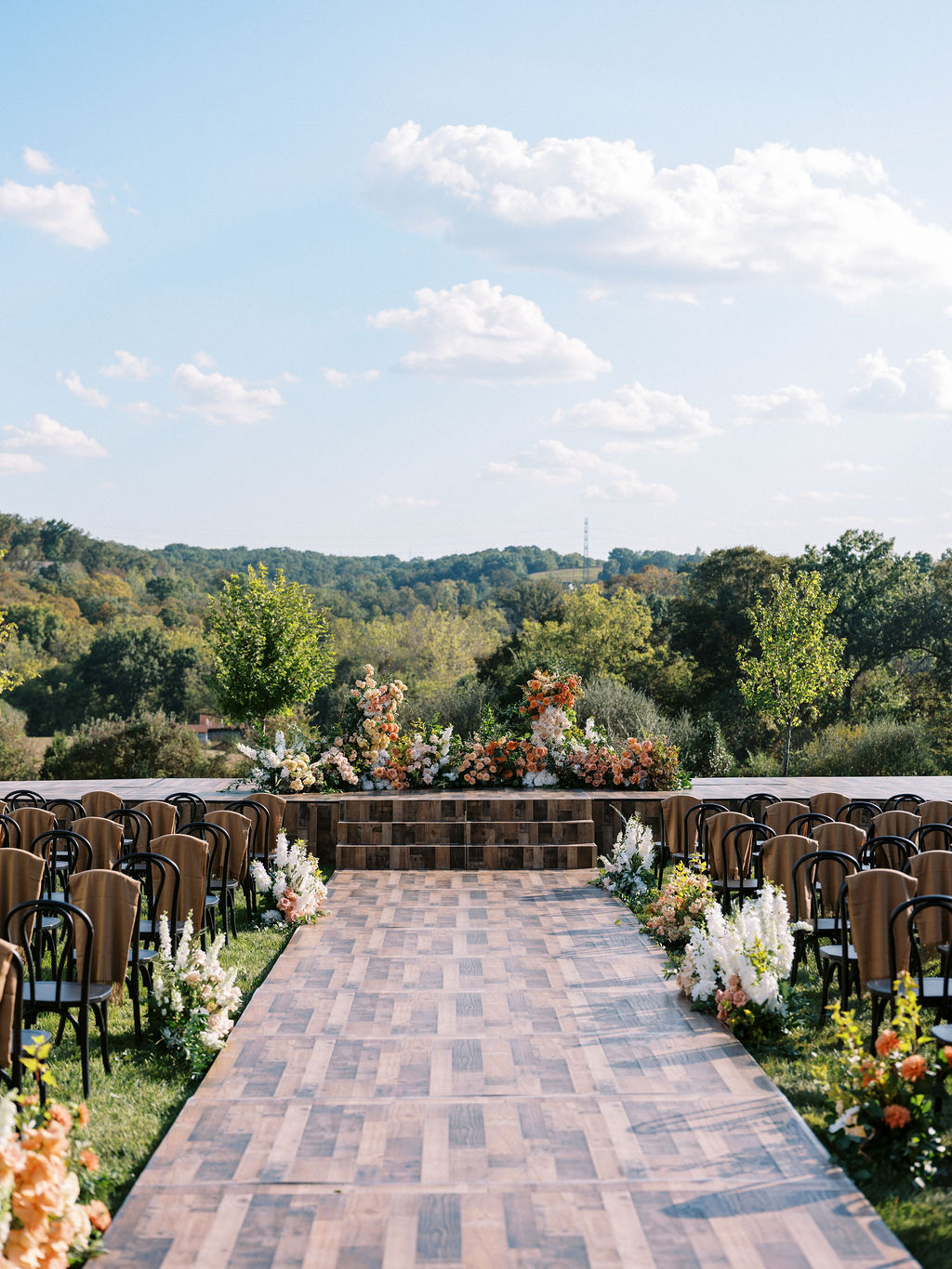 Outdoor wedding ceremony setup with chairs, floral arrangements, and a scenic landscape background under a partly cloudy sky.
