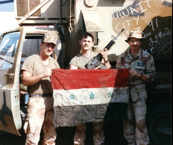 Three soldiers holding a flag with red, white, and black stripes and blue stars, standing outdoors near a vehicle.