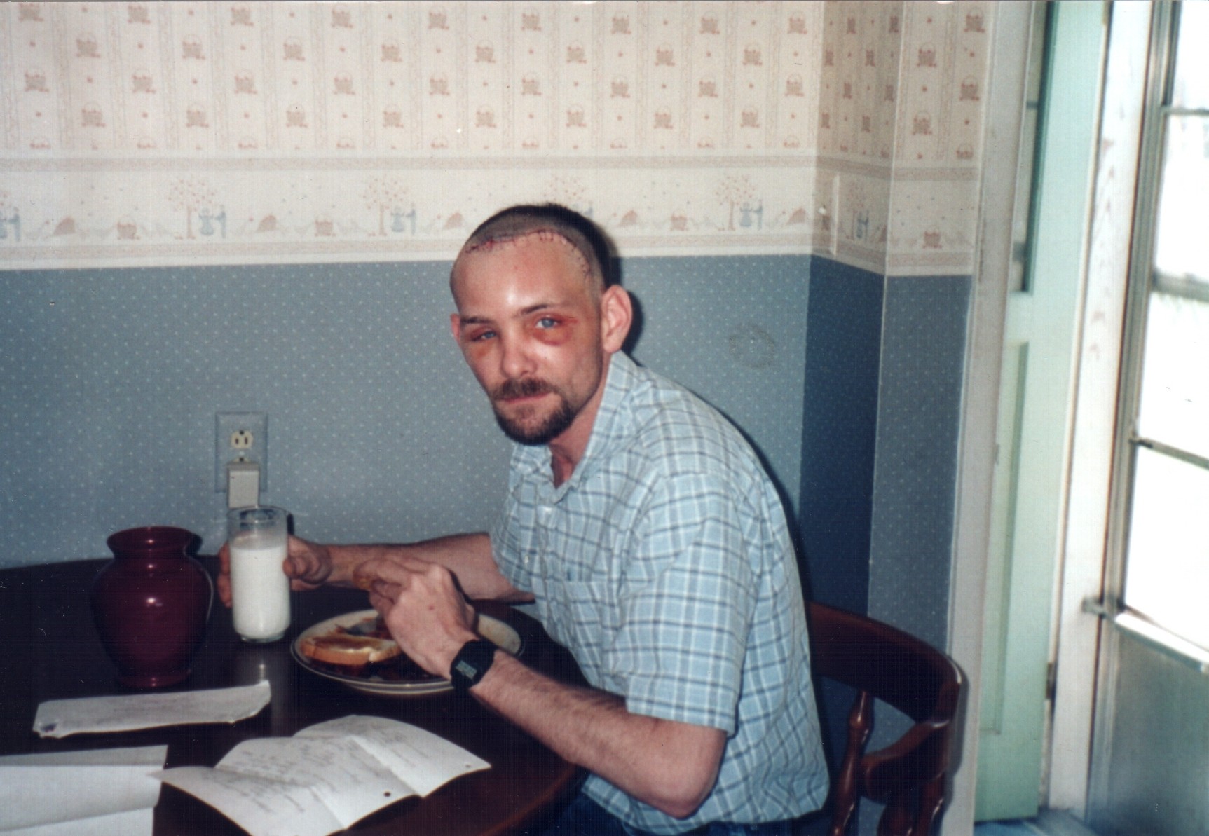 Man sitting at a table with a plate, a glass of milk, and a book, near a window with sunlight.