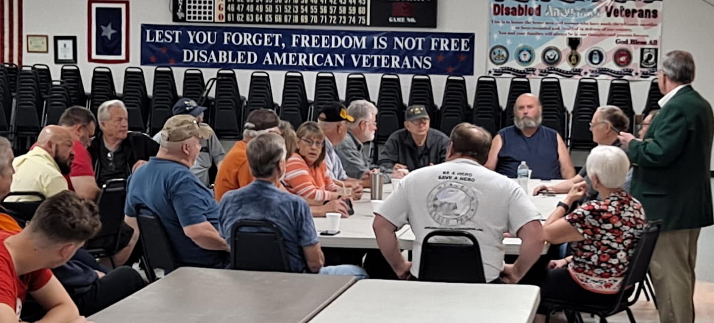 Group of people seated around tables in a room with banners honoring disabled American veterans, listening to a speaker.