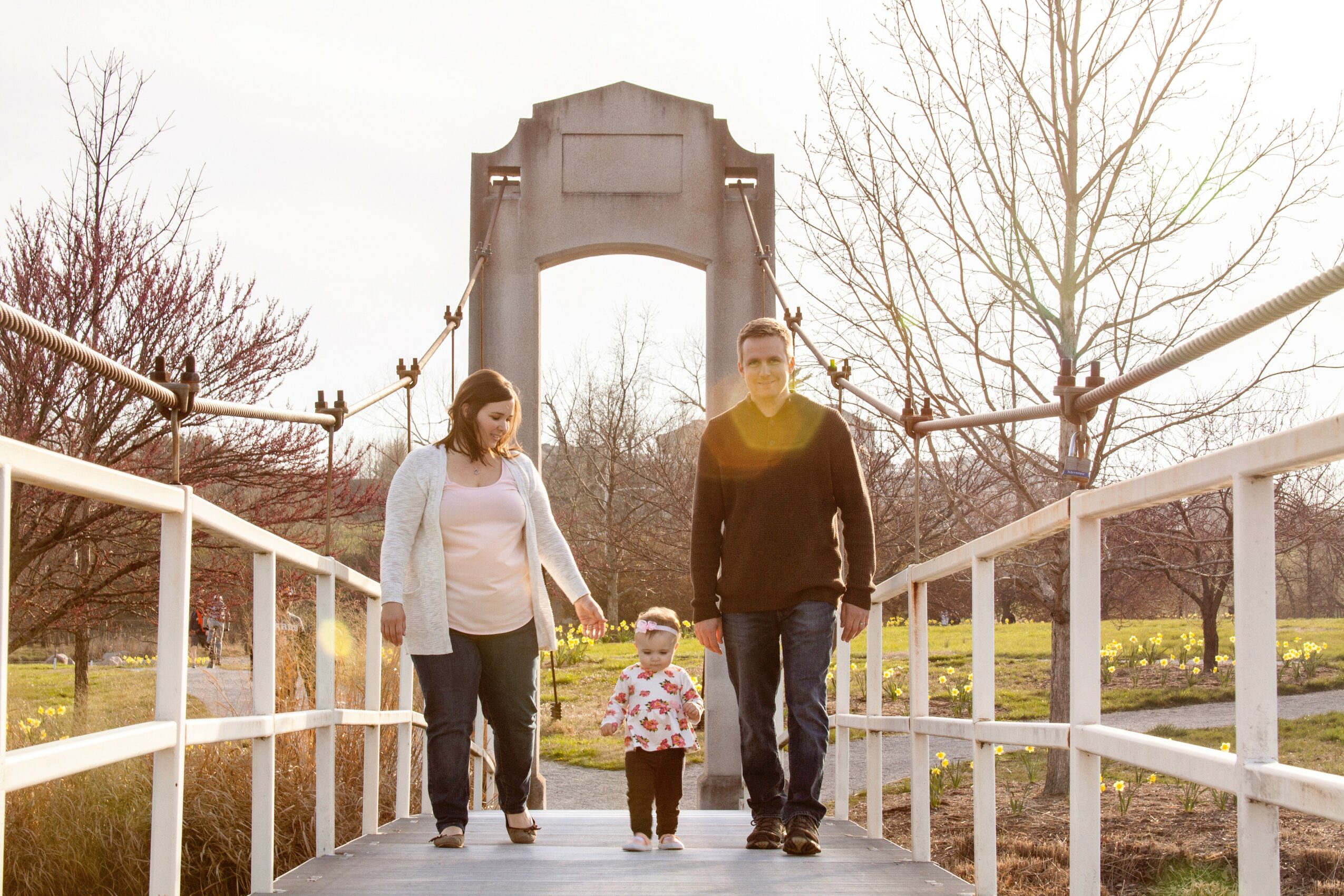 A family of three walking on a bridge with trees and a structure in the background.