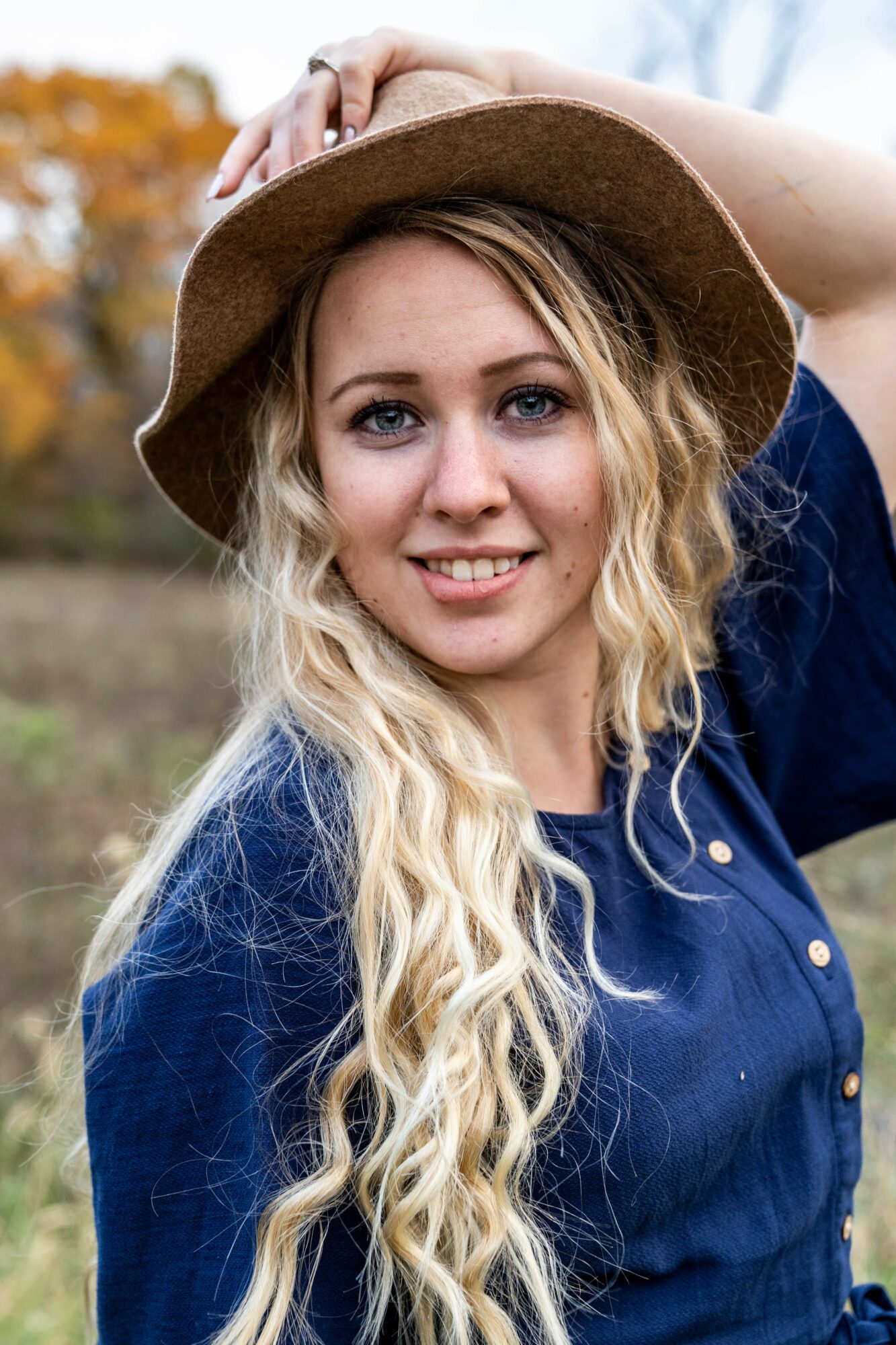 Young woman with long blonde hair wearing a wide-brimmed hat and blue shirt outdoors, smiling.