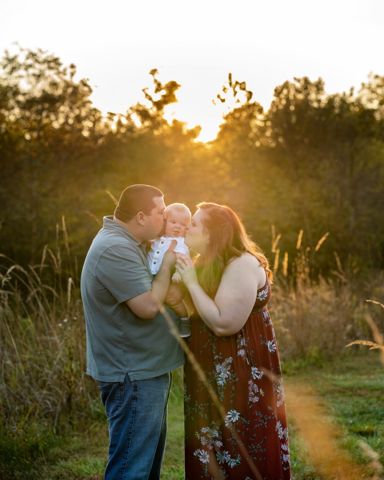 Family of three outdoors at sunset, with trees and grass in background, holding a baby and smiling.