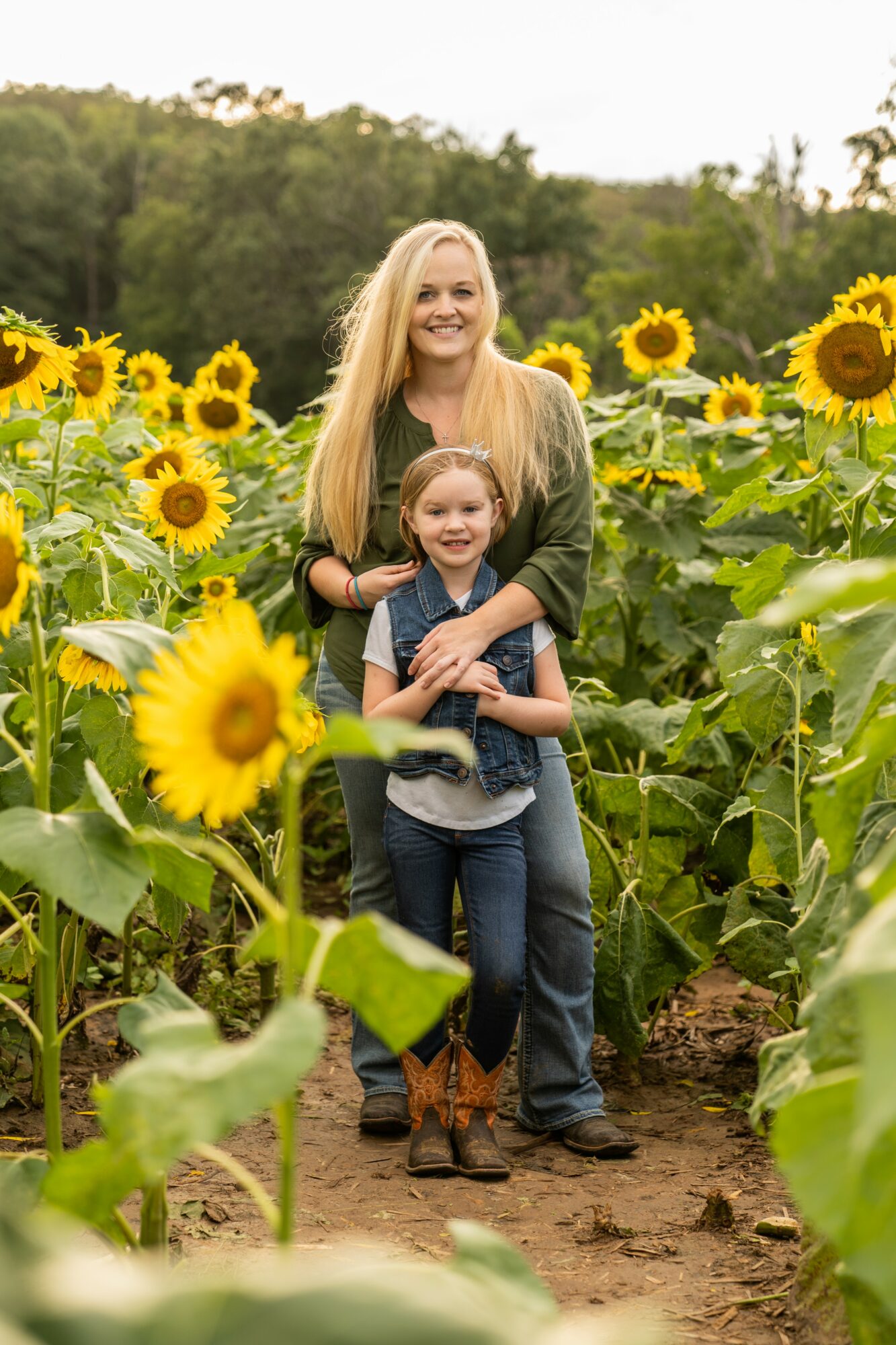 A woman and a girl stand in a sunflower field, smiling, with green trees in the background.