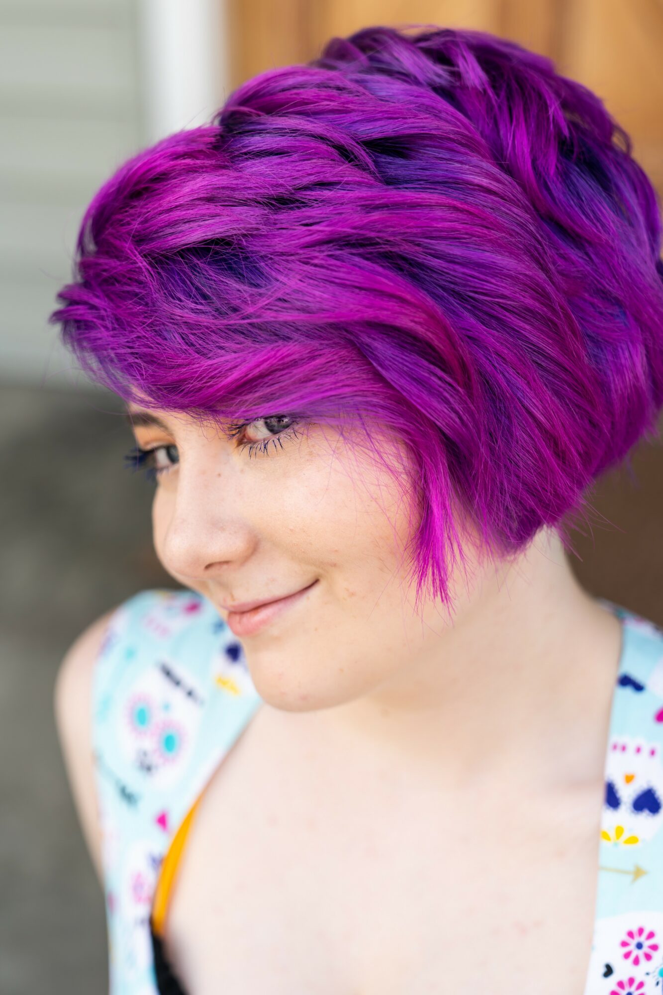 Young woman with short, vibrant purple hair, smiling gently, wearing a colorful patterned top, outdoors.