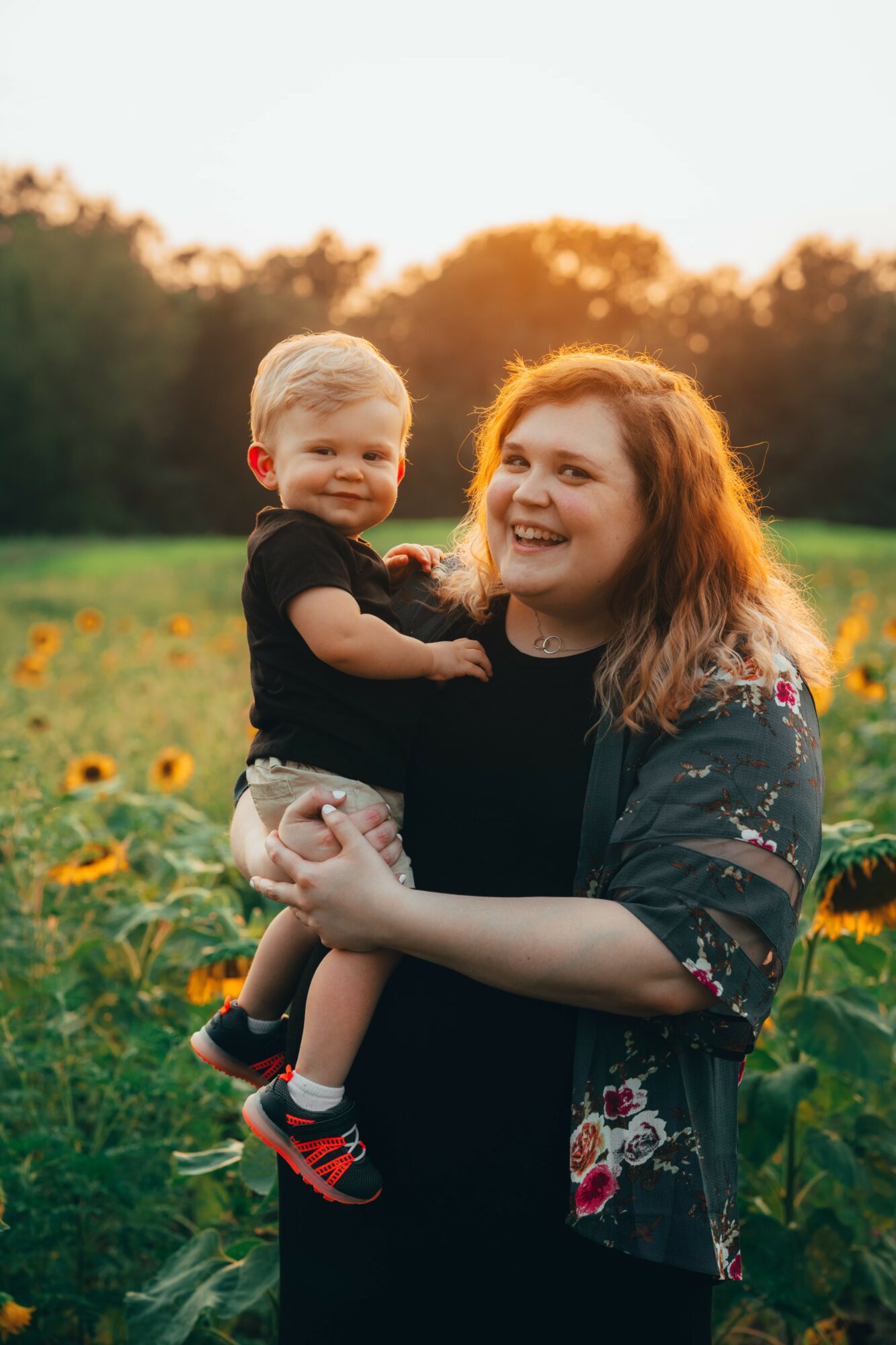 Woman holding a smiling child in a sunflower field during sunset, both looking happy. Bright sunlight in background.