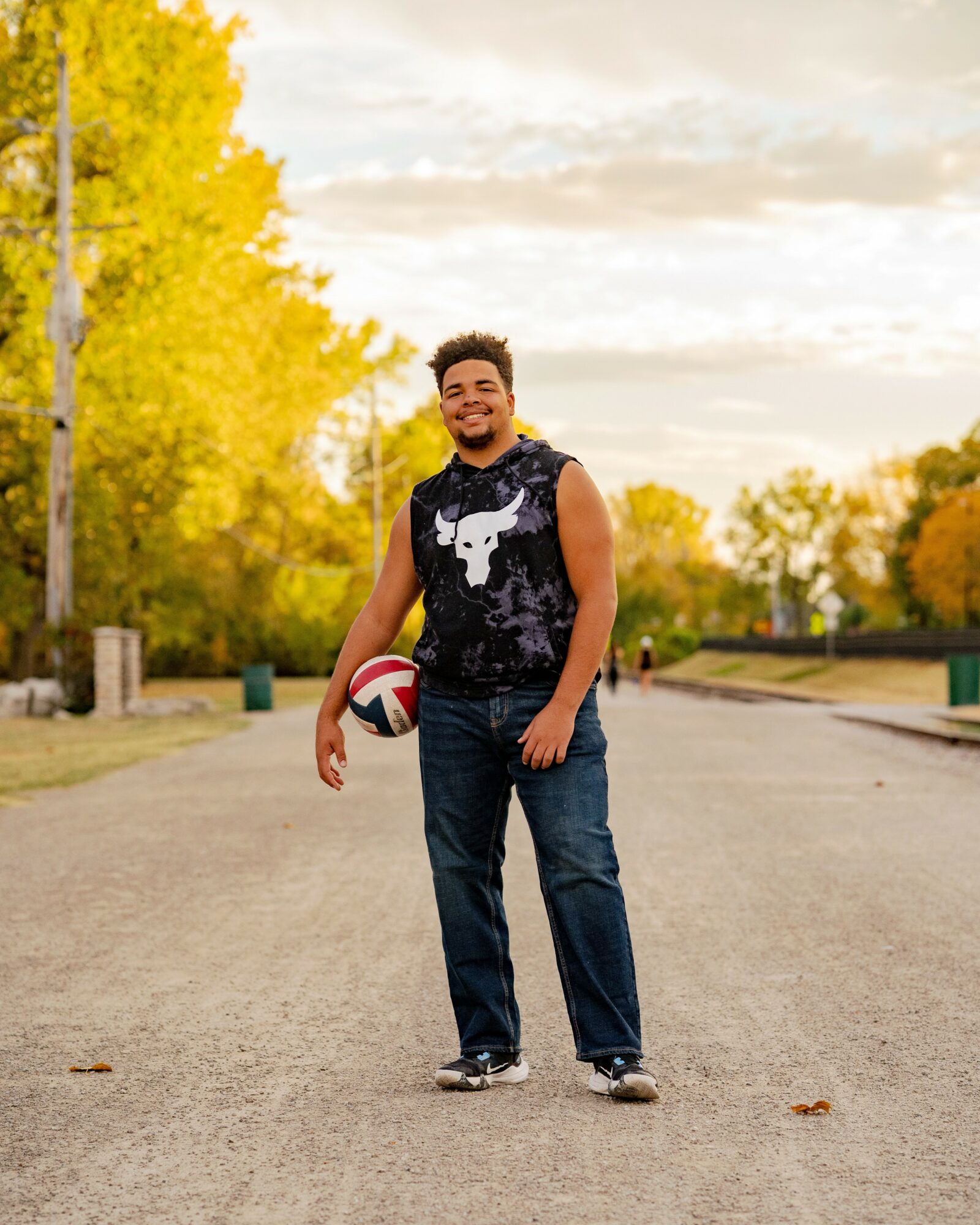 Young man standing on a paved path holding a volleyball, smiling, with trees and a cloudy sky in the background.