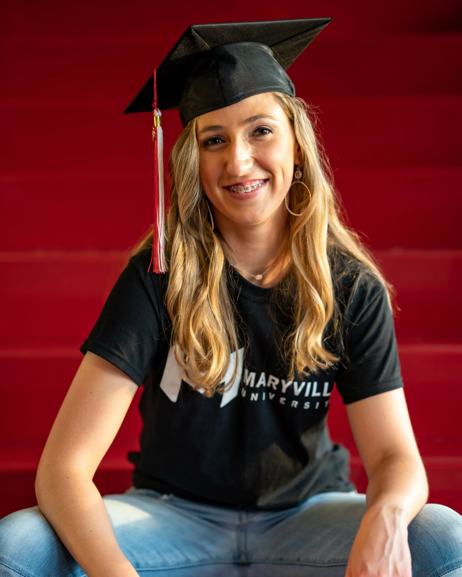Young woman in graduation cap and black shirt smiling, sitting against a red background.