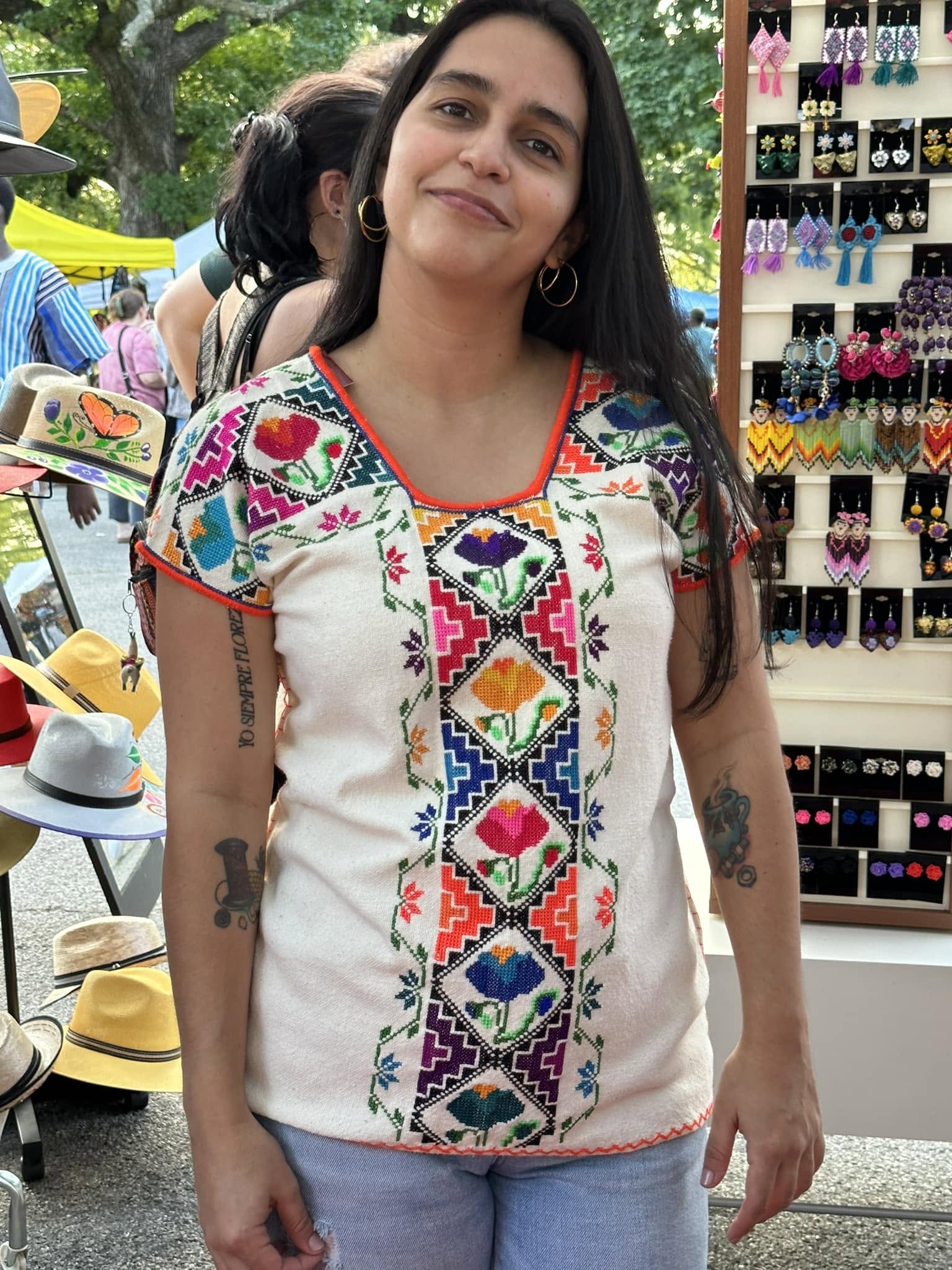 Woman with long dark hair wearing a colorful embroidered blouse, standing outdoors near a display of hats and accessories.