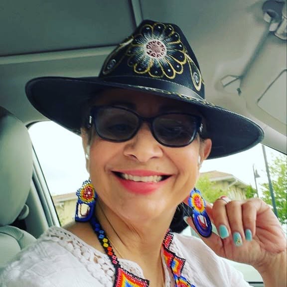 Smiling woman wearing a wide-brimmed hat, sunglasses, colorful earrings, and a white top inside a vehicle.