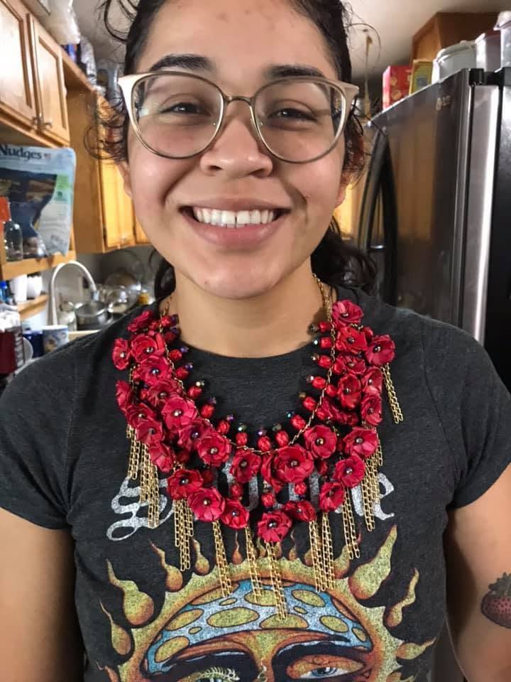 Smiling person with glasses and a red floral necklace in a kitchen, wearing a dark T-shirt with a colorful design.