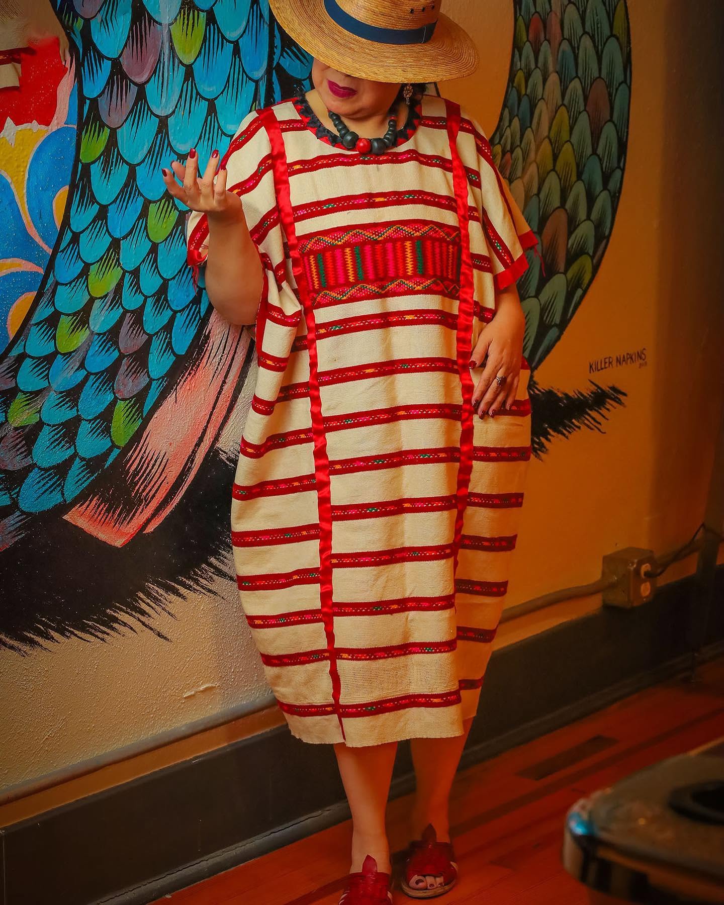 Person in a striped dress with red accents, wearing a wide-brimmed hat, standing indoors near colorful wall art.