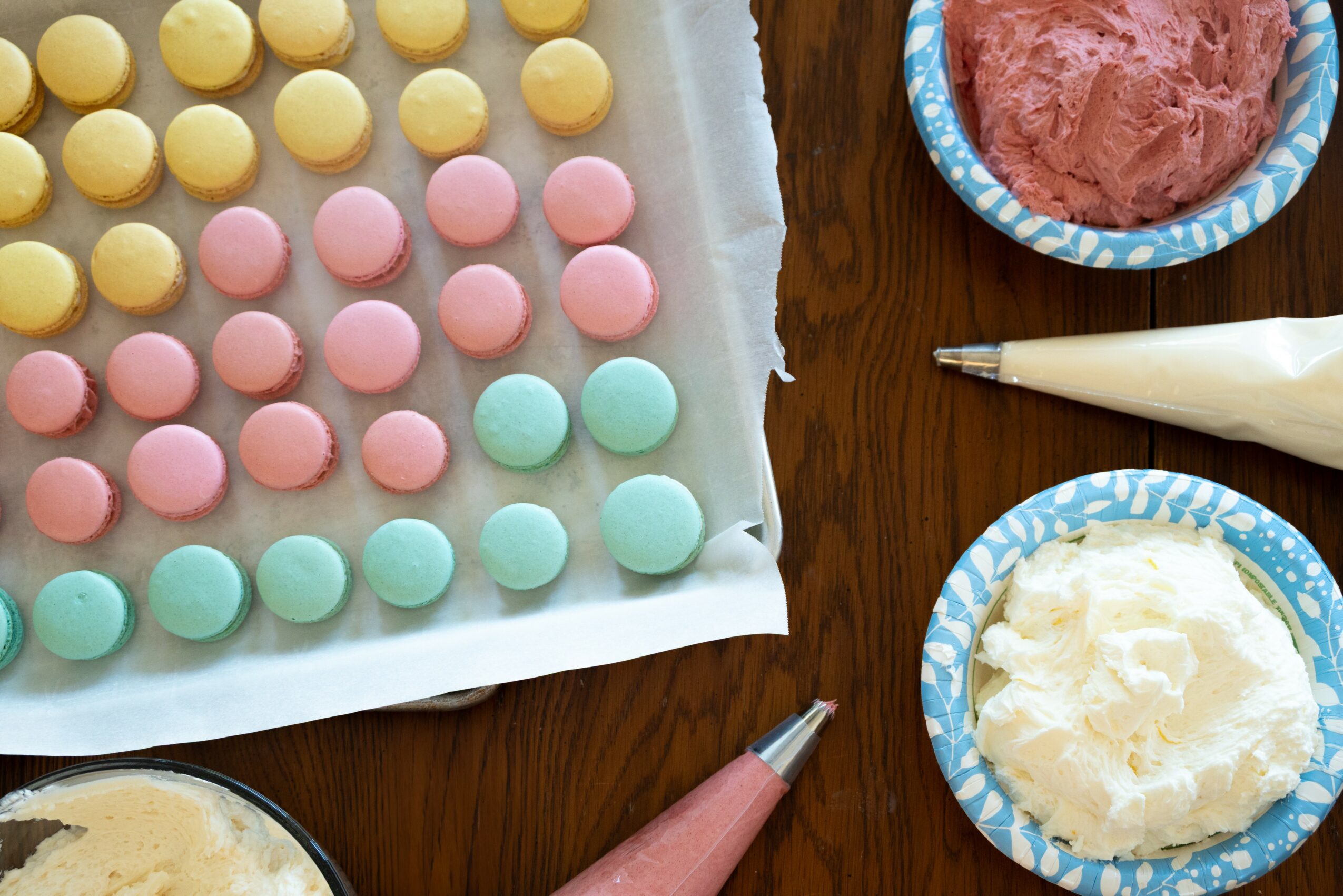 Colorful round candies on parchment paper, pink, yellow, and blue, with frosting and piping bags on a wooden surface.