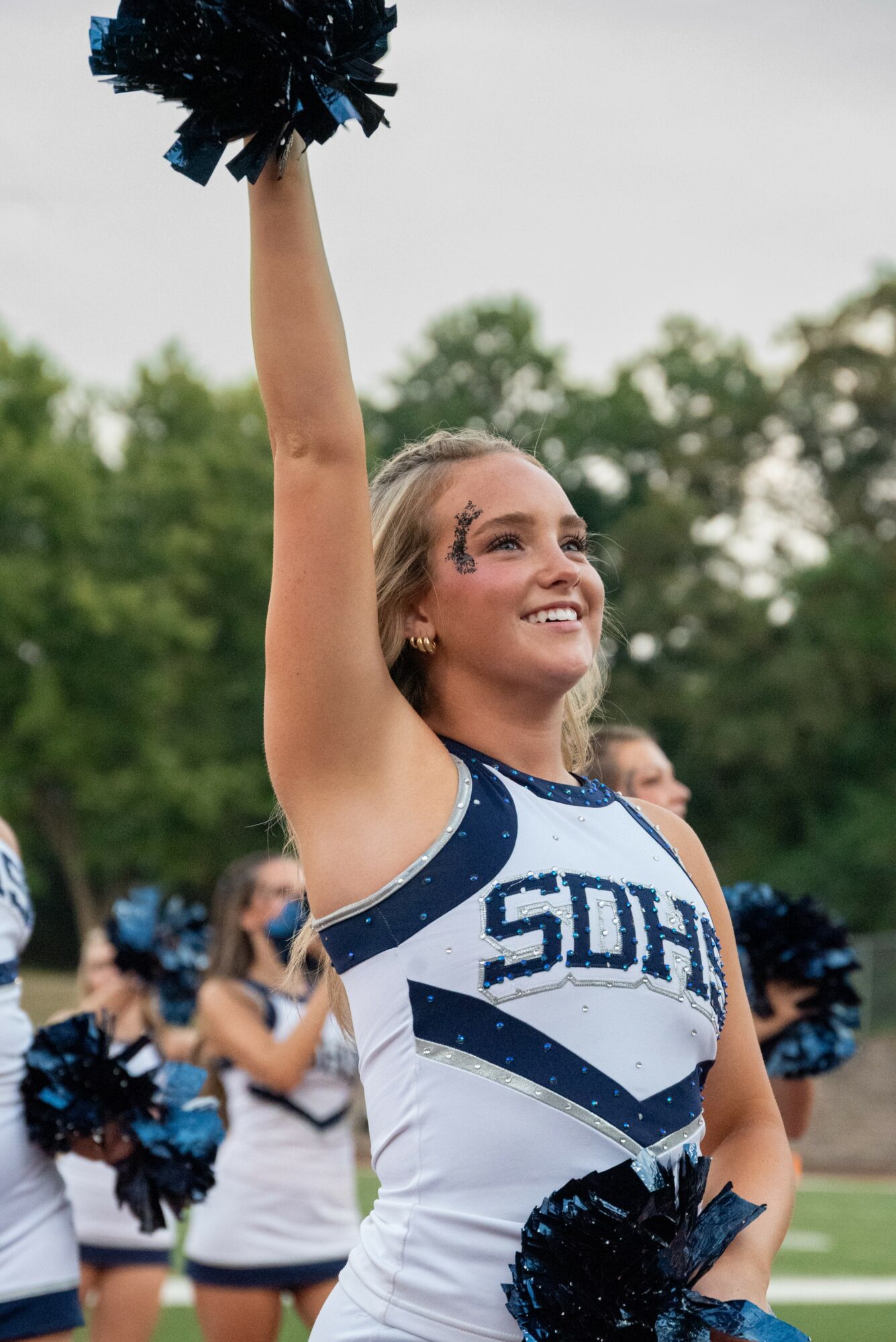 Cheerleader smiling and raising pom-pom in outdoor sports field, trees in background.