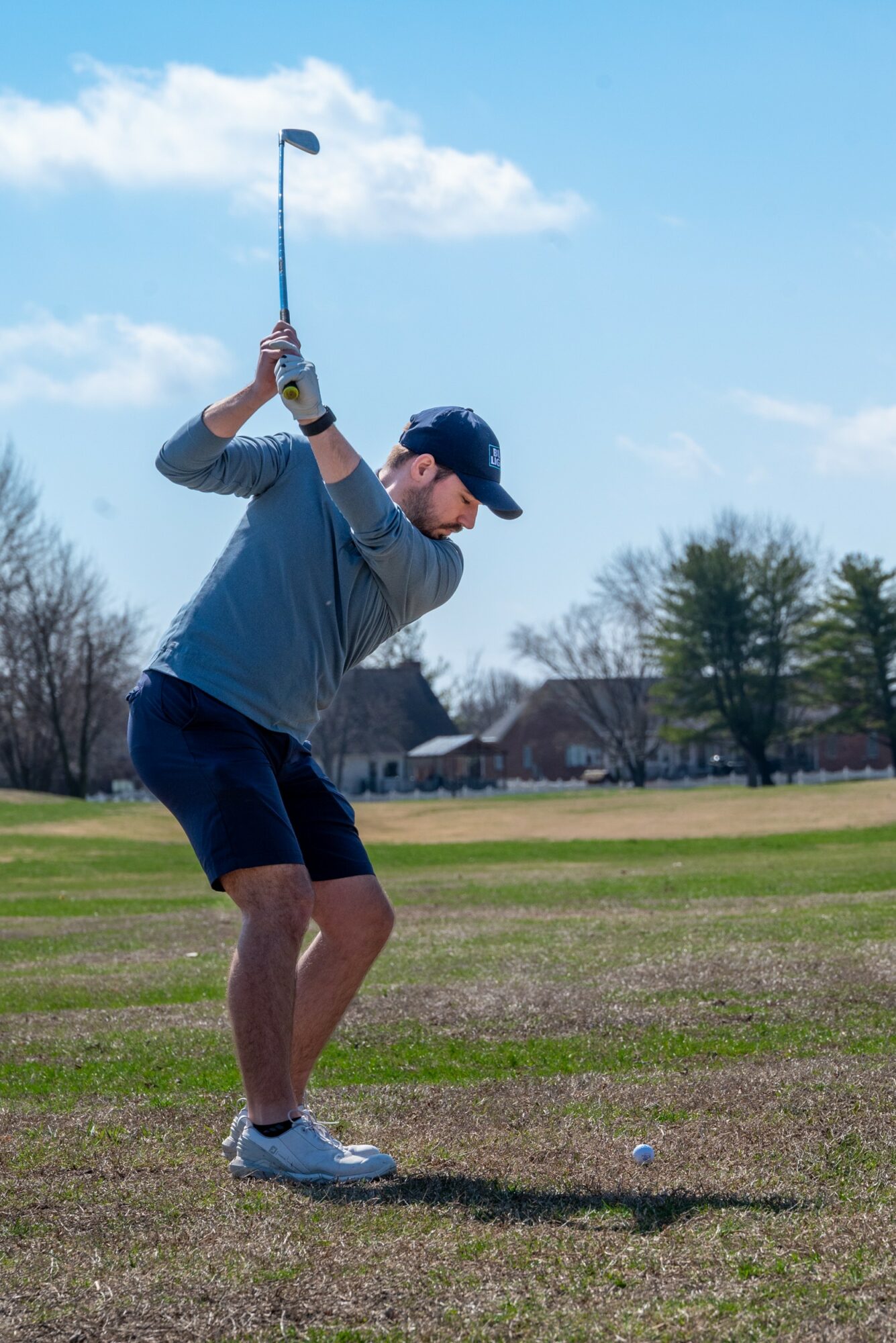 Person preparing to hit golf ball on course, wearing cap, gray shirt, and shorts, with trees and houses in background.