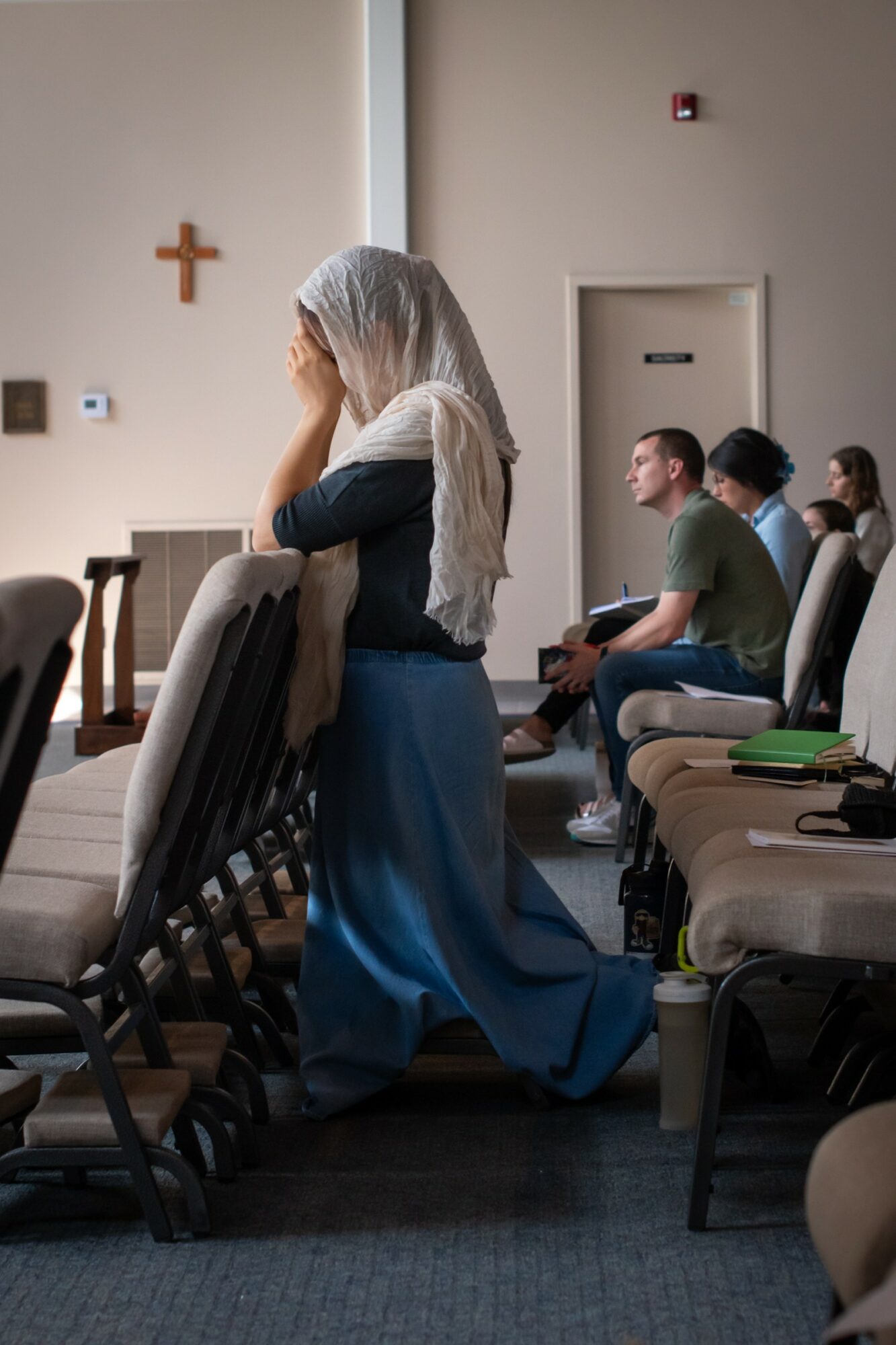 A woman with a head covering kneels in prayer in a church, with people seated behind her. Cross on wall.