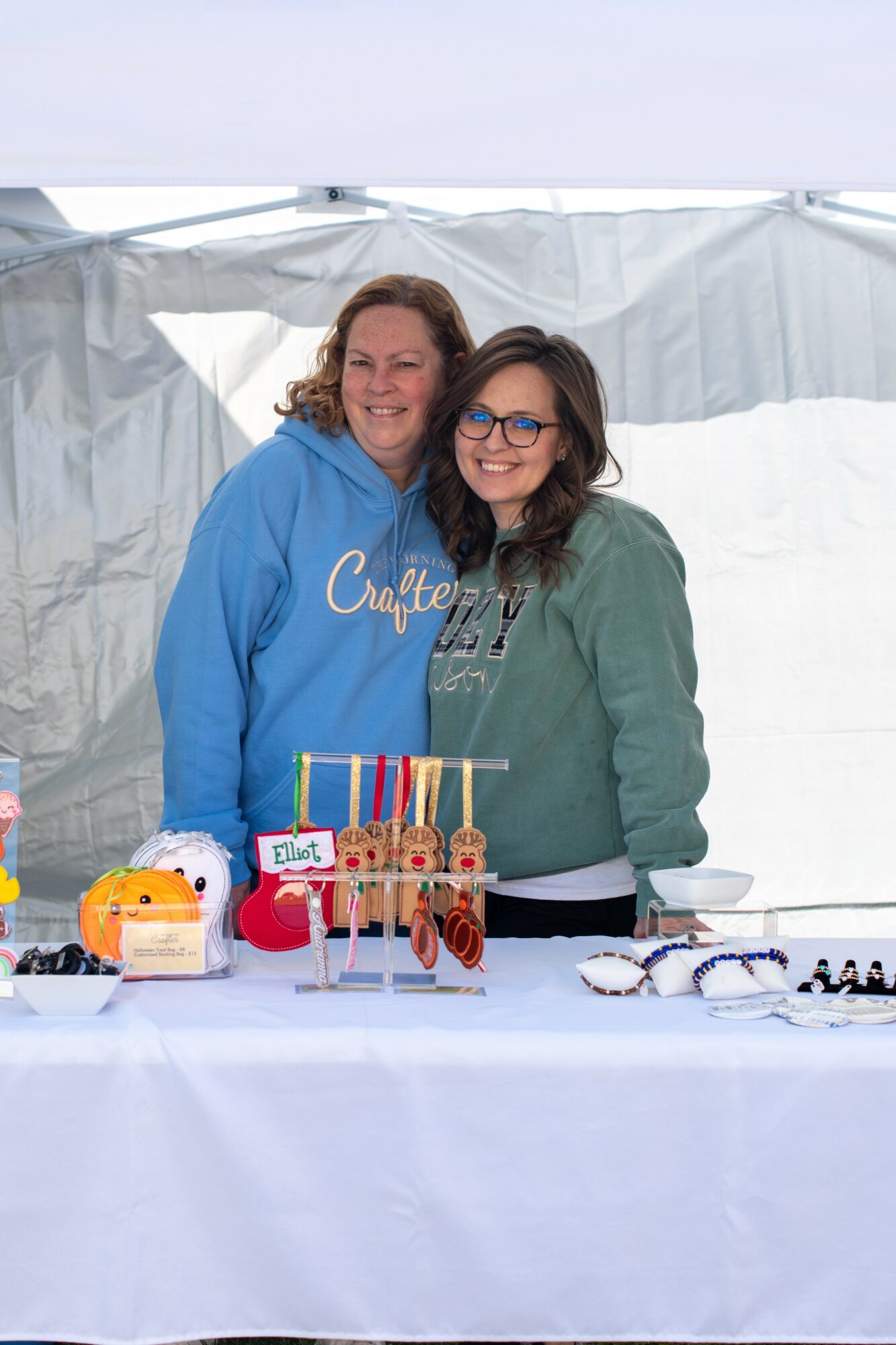 Two women smiling behind a table with crafts and jewelry, inside a tent, one wearing a blue hoodie and glasses.