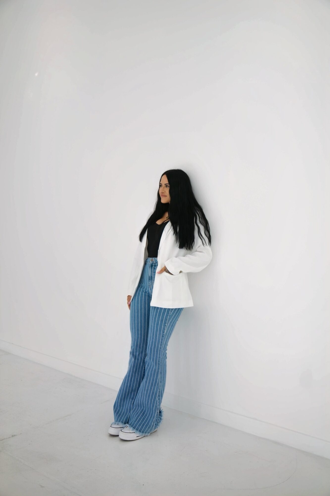 Woman with long dark hair wearing a white blazer, black top, and striped blue pants, standing against a plain white wall.