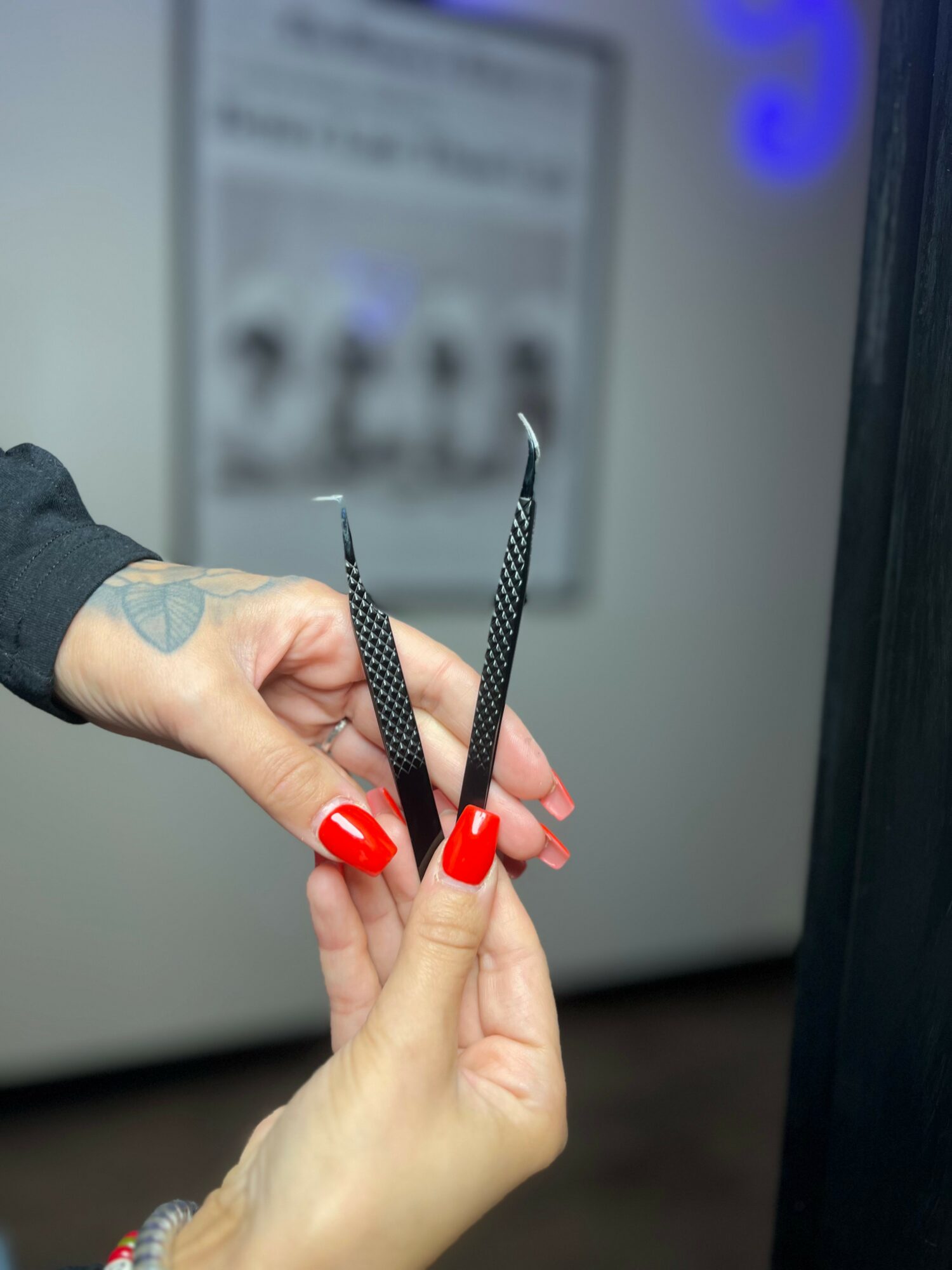 Two hands holding black and white striped tweezers, one with red nail polish, blurred background with a poster or sign.