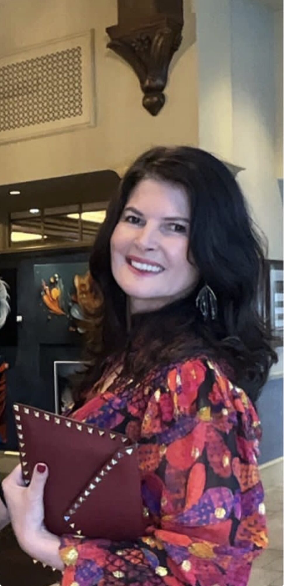 Woman with dark hair smiling, holding a red clutch, wearing a colorful patterned top, in an indoor setting.