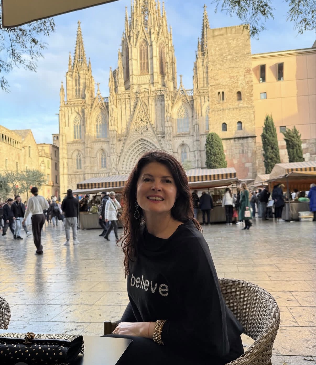 Woman sitting outdoors in front of a historic cathedral, smiling, with market stalls and people in the background.
