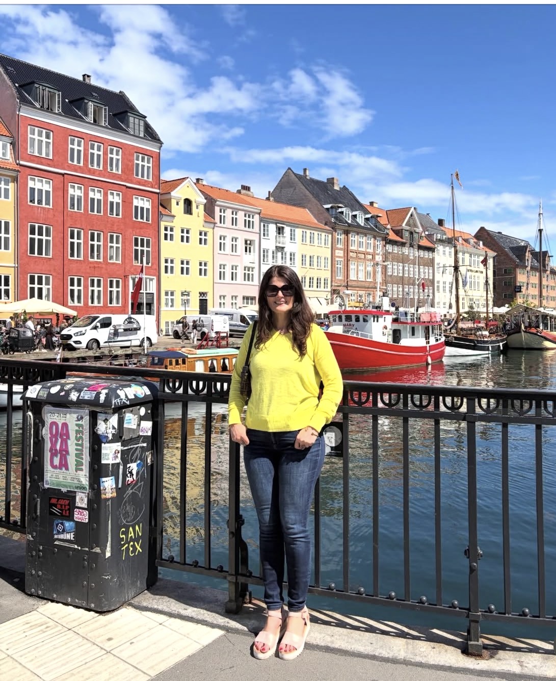 Woman in yellow jacket and sunglasses standing by canal with colorful buildings and boats in background.