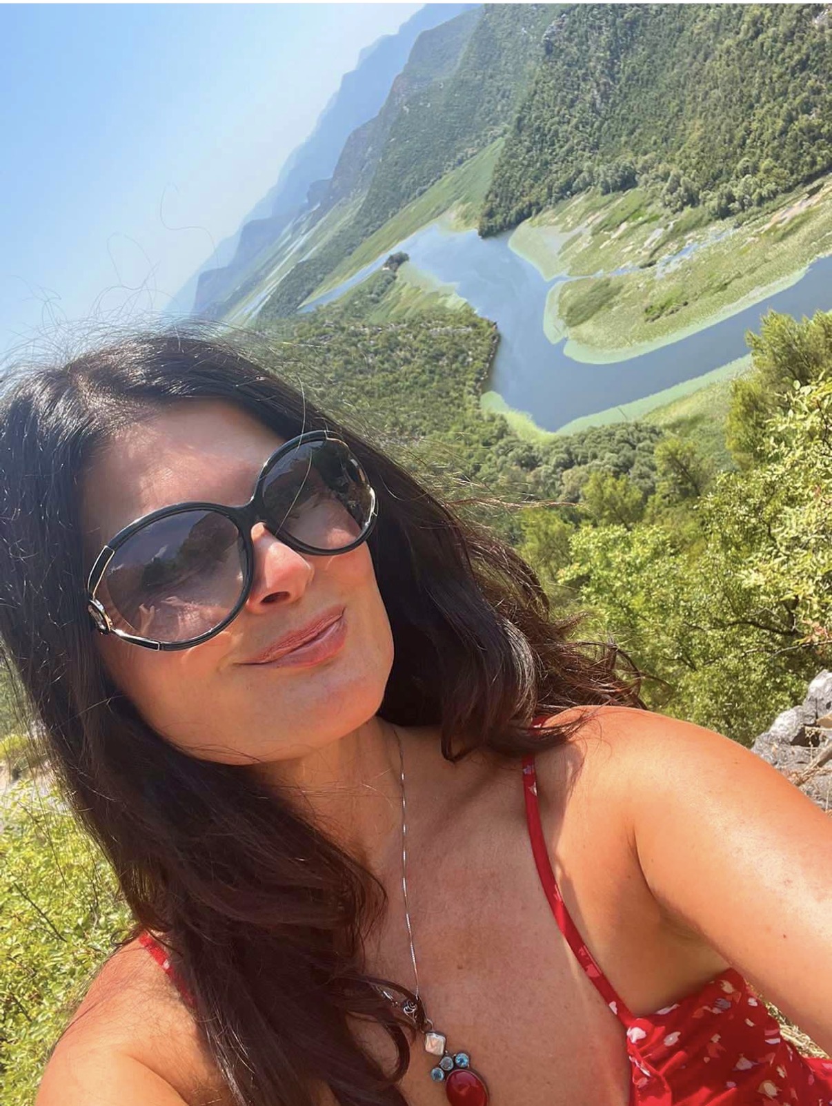 Woman with sunglasses taking a selfie overlooking a winding river and green landscape, sunny day.
