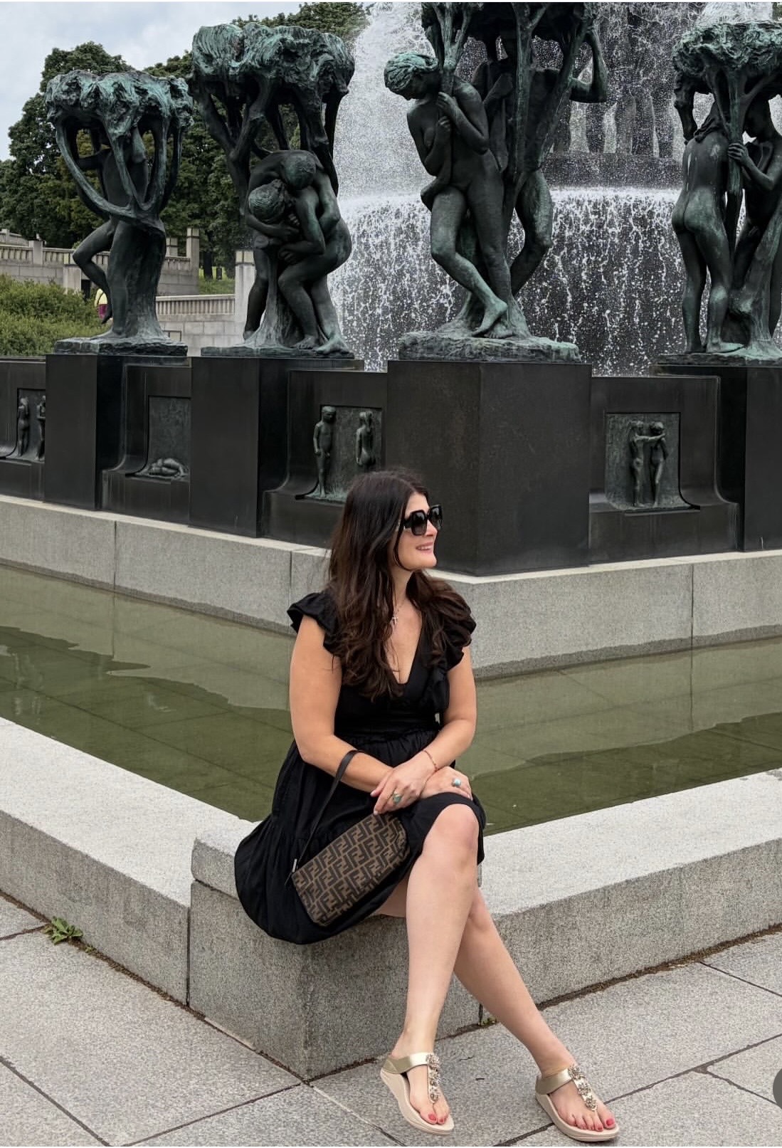 Woman sitting on a stone ledge near a fountain with bronze sculptures in the background, wearing sunglasses and a black dress.
