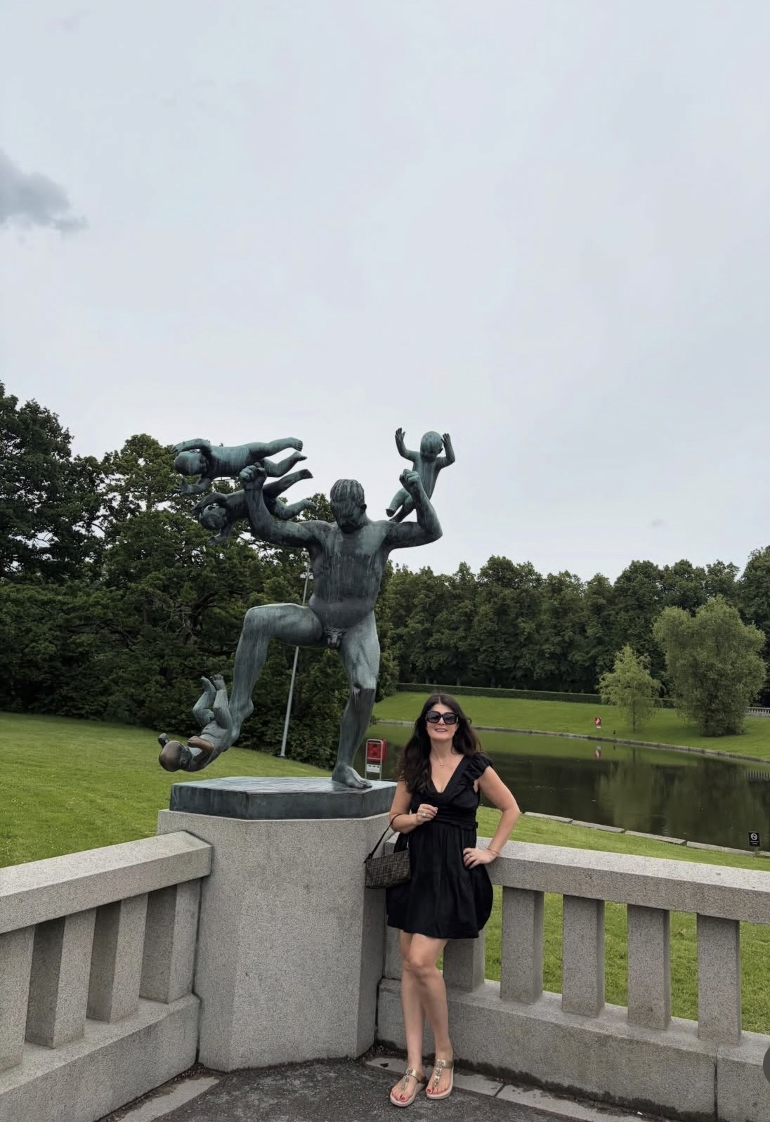 Woman in black dress and sunglasses standing on a bridge with a large sculpture of a person and three small figures behind her, trees and a pond in the background.