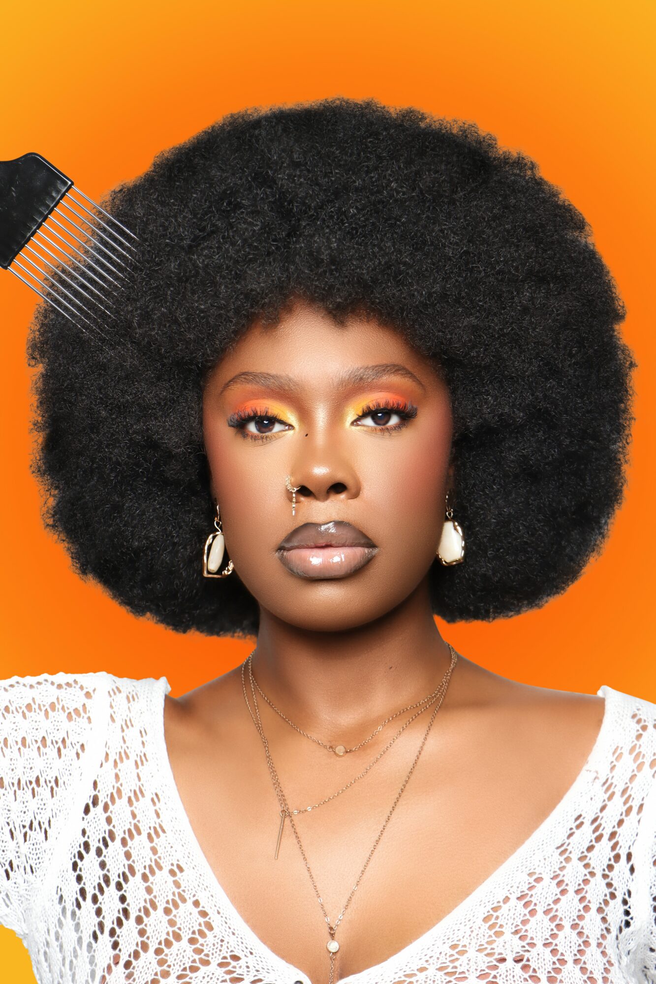 Woman with large afro hairstyle, wearing earrings, a white crochet top, and layered necklaces, against an orange background.