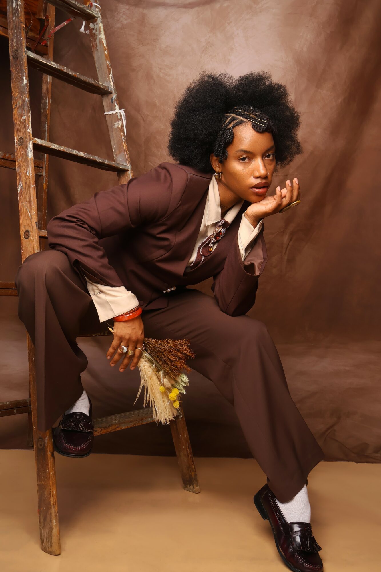 Woman with natural hair in a brown suit, sitting on a wooden chair, holding a small bouquet of flowers, against a brown backdrop.