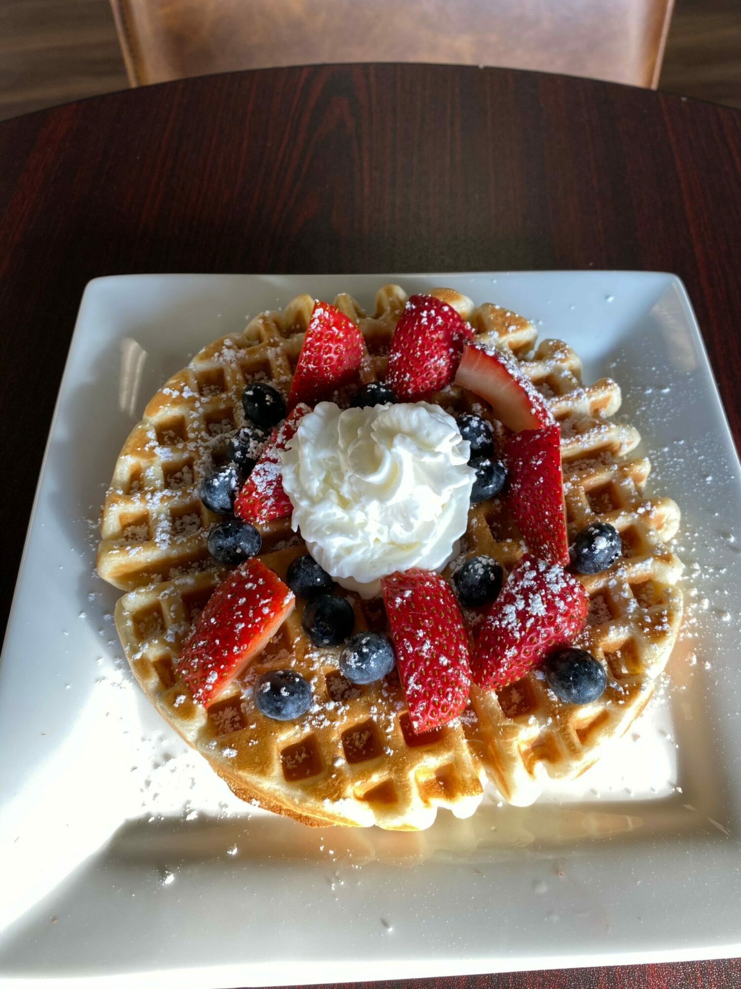 Waffle topped with strawberries, blueberries, whipped cream, and powdered sugar on a white plate.