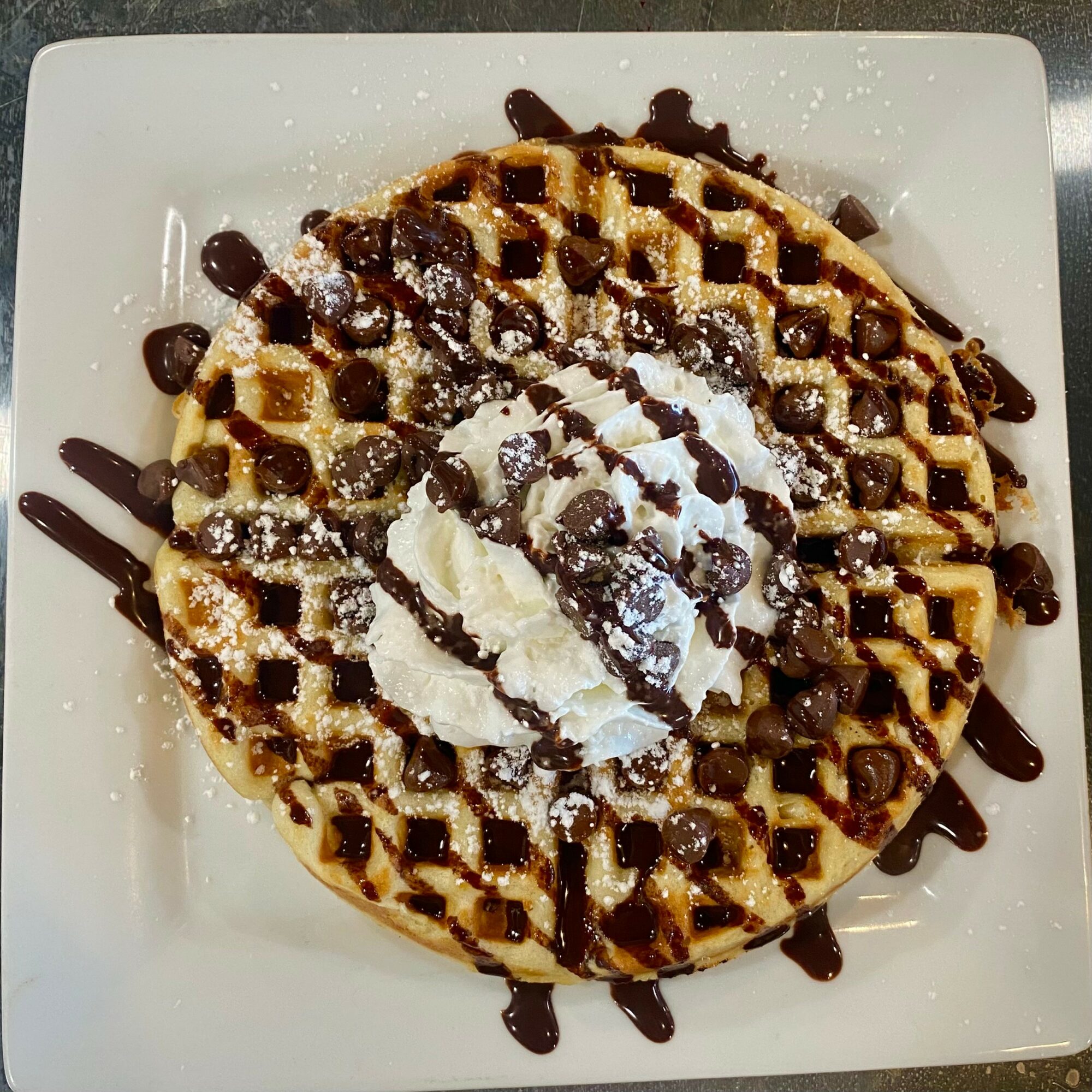 Round waffle topped with whipped cream, chocolate drizzle, and powdered sugar on a white plate.