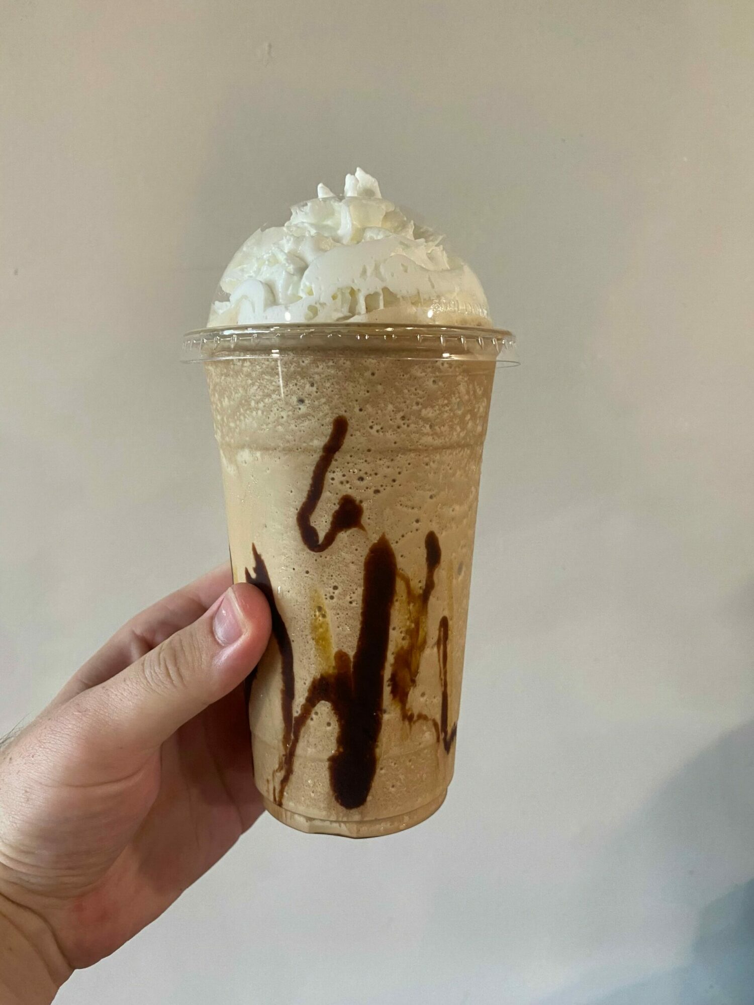 Hand holding a blended coffee drink with whipped cream and chocolate syrup, against a plain background.