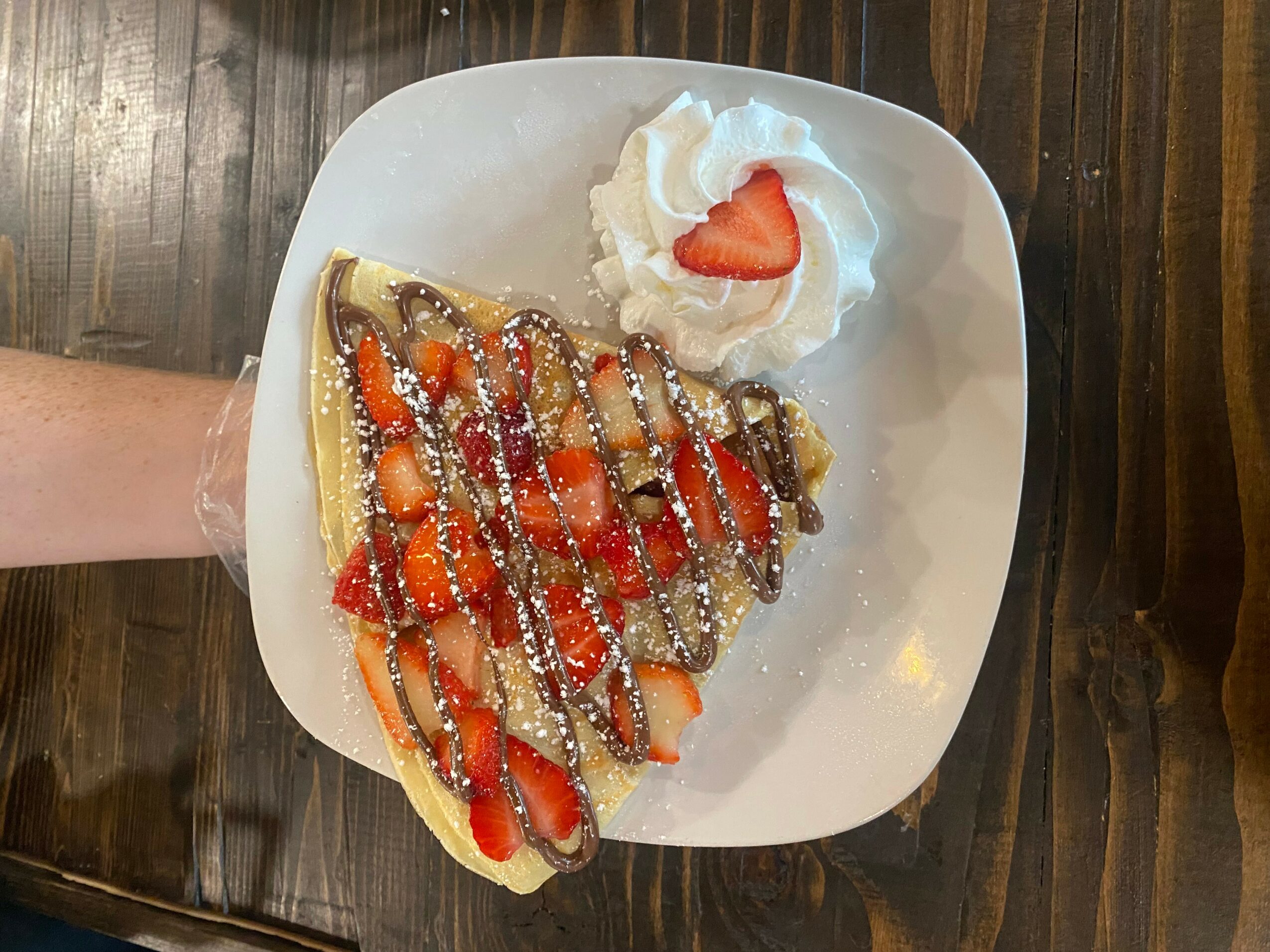 A plate with a crepe topped with strawberries, powdered sugar, and chocolate drizzle, served with whipped cream and a strawberry slice.