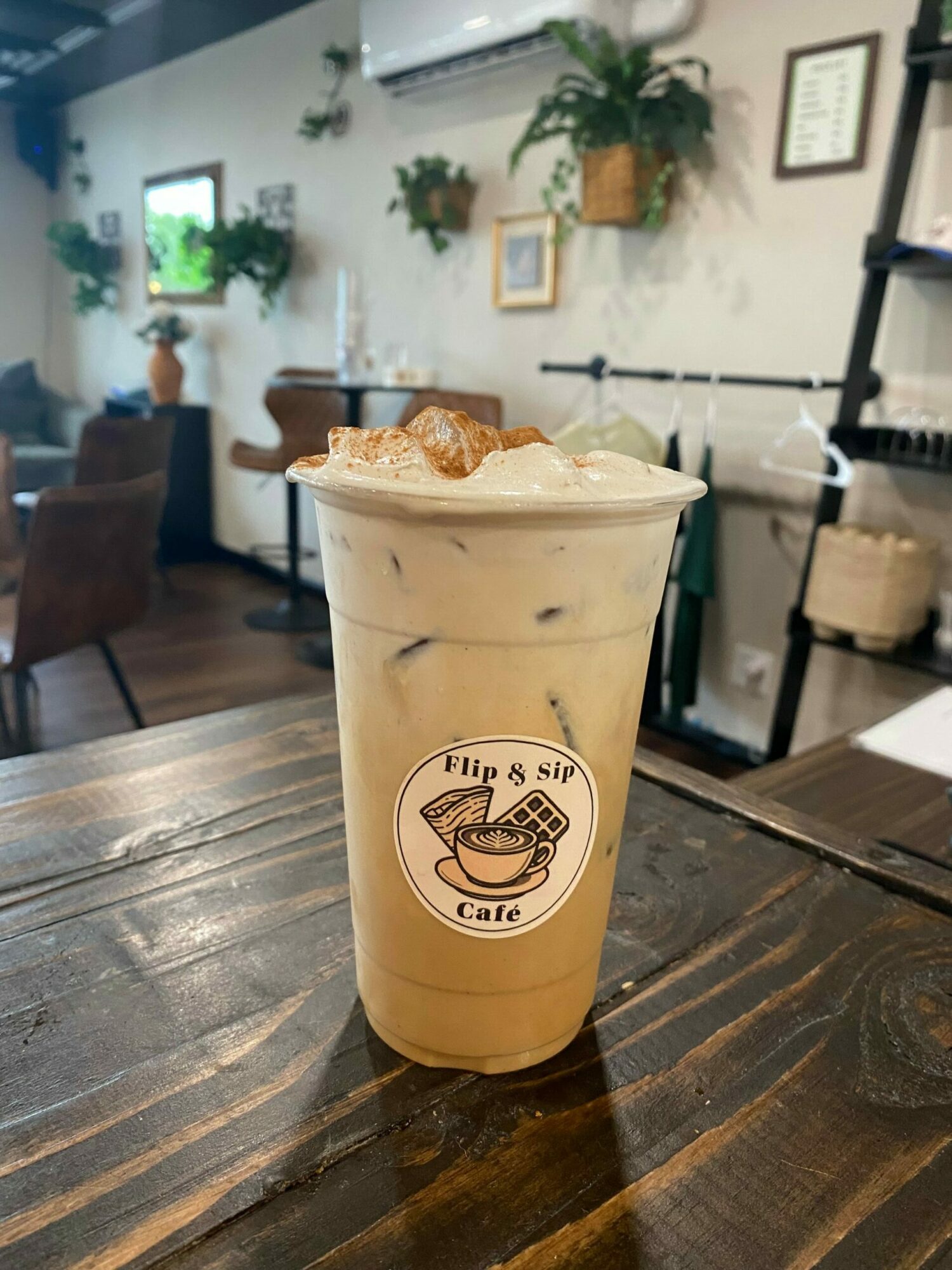 Iced coffee in a clear plastic cup with a logo, topped with whipped cream and caramel drizzle, on a wooden table.