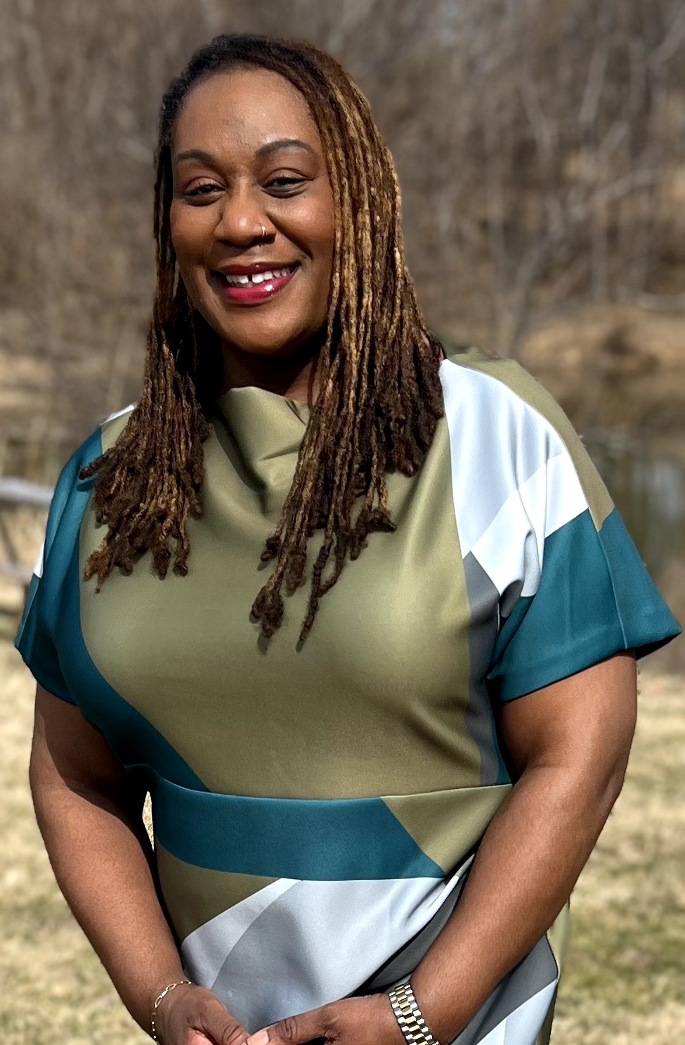 Smiling woman with long dreadlocks outdoors, wearing a colorful top, standing in natural setting.