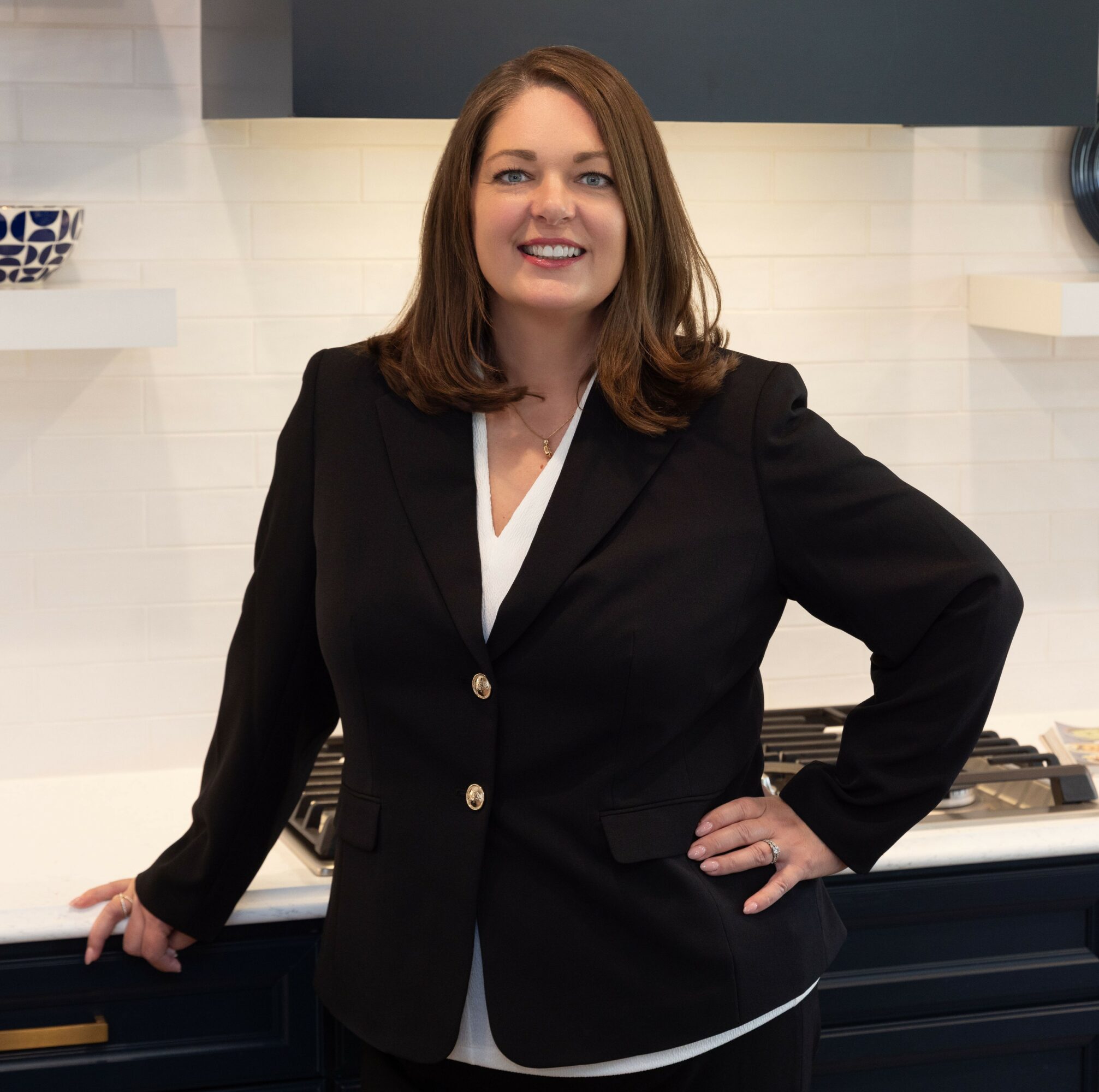Woman with shoulder-length brown hair smiling, wearing a black blazer and white blouse, standing in a modern kitchen.