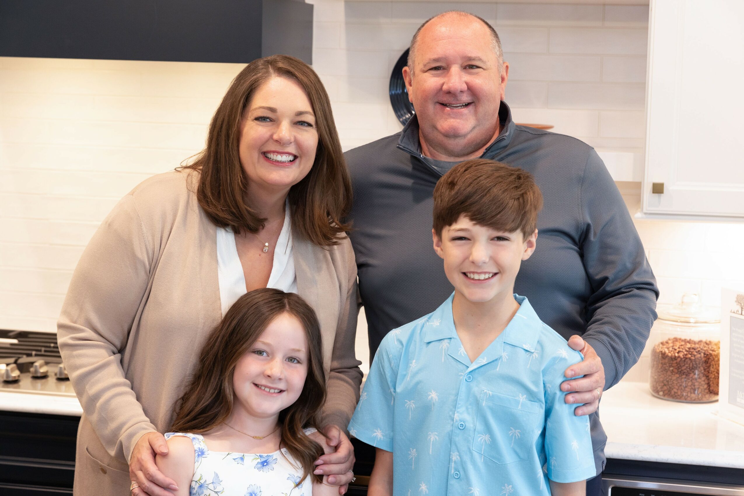 Four people smiling in a kitchen, two adults and two children, standing close together.