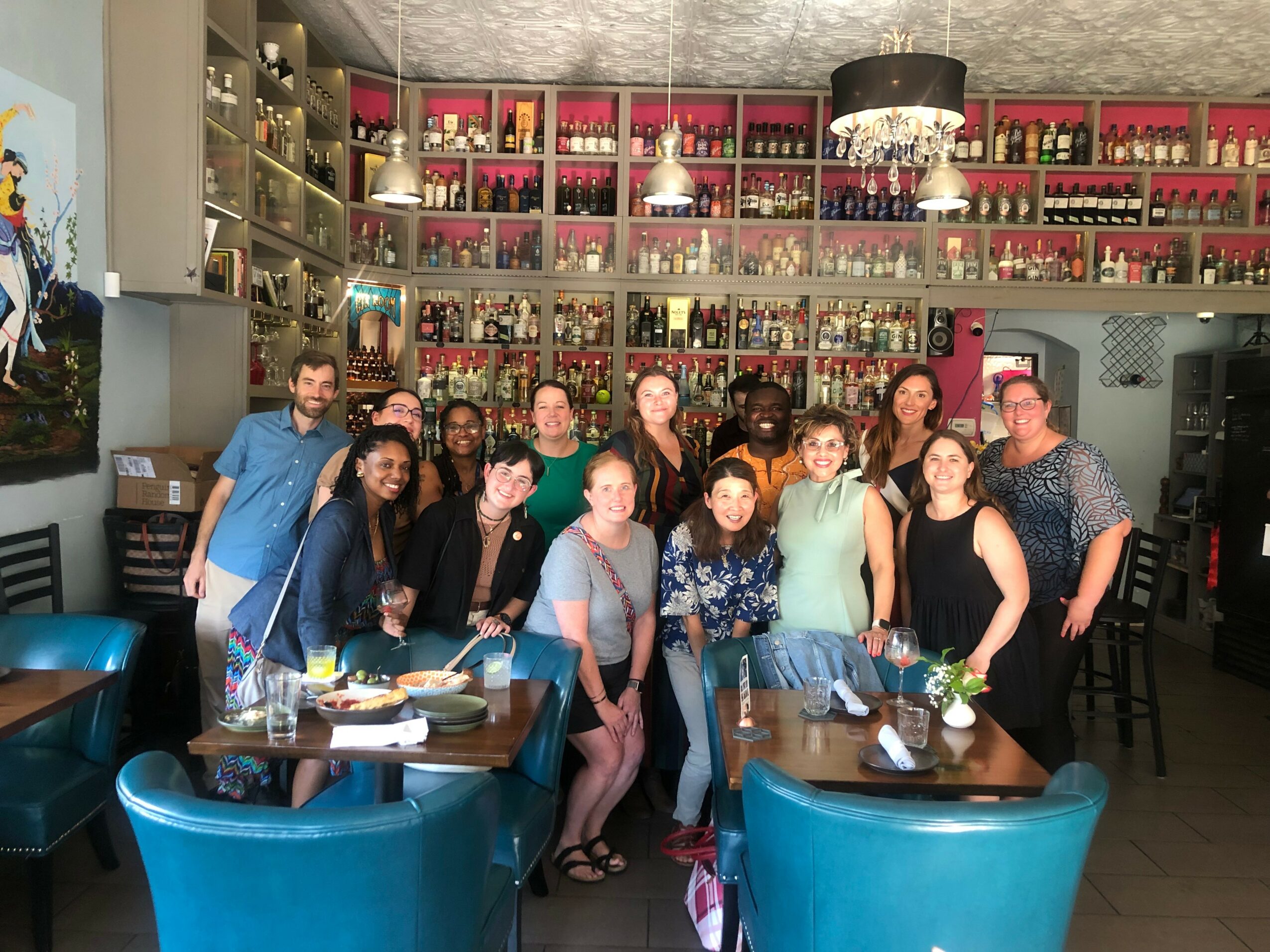 Group of people gathered in a bar or restaurant with shelves of bottles behind them, some sitting and some standing, smiling.