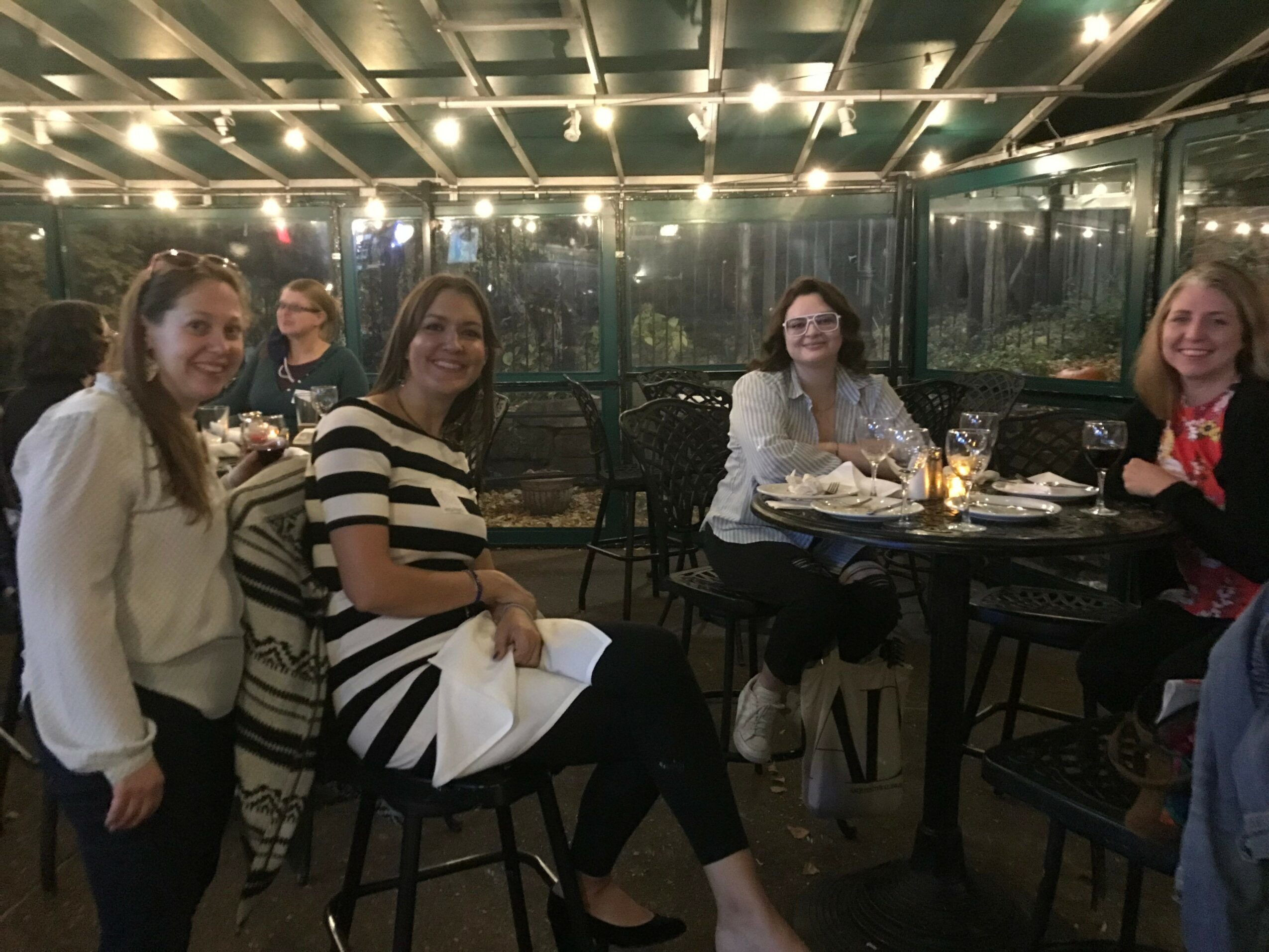 Group of five women sitting and standing at a table in a restaurant or cafe, smiling, with glasses and plates on the table.
