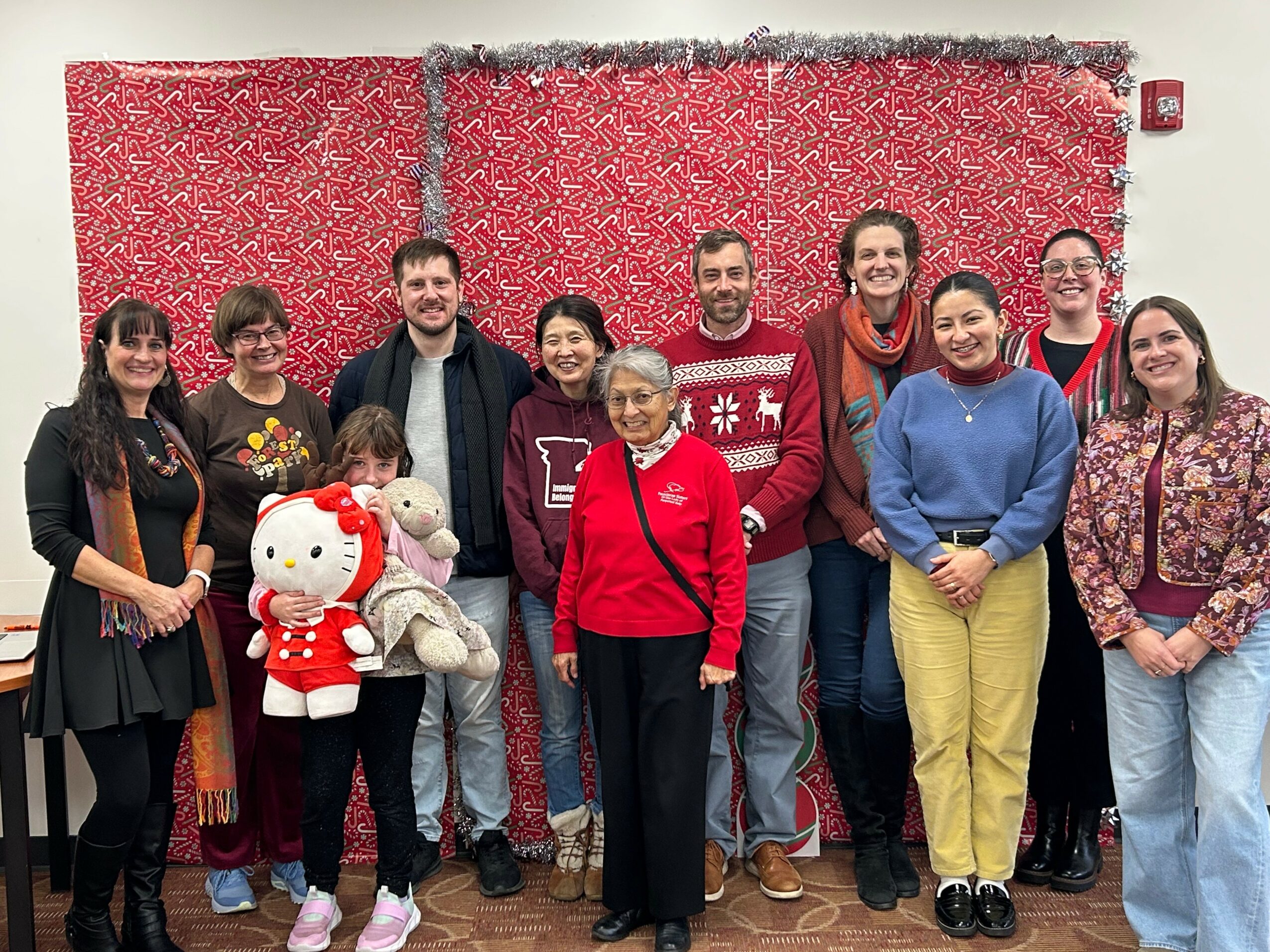 Group of ten people standing indoors in front of a red patterned wall, smiling, with a Hello Kitty plush toy, diverse ages and styles.