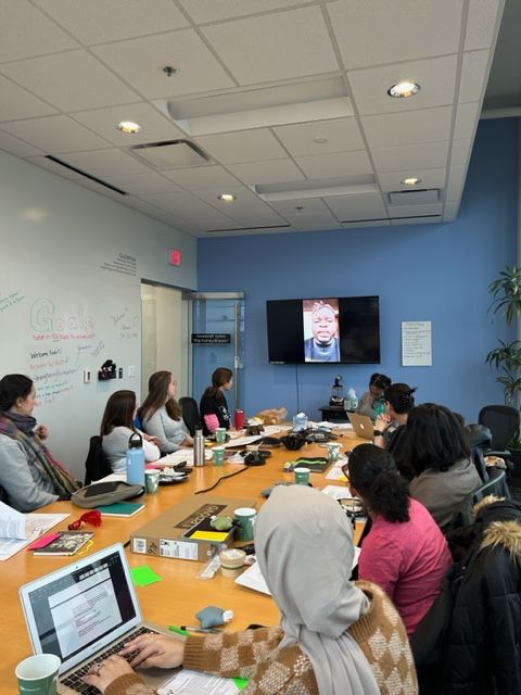People seated around a conference table in a room with a blue wall, a whiteboard, and a television screen, engaging in a meeting.