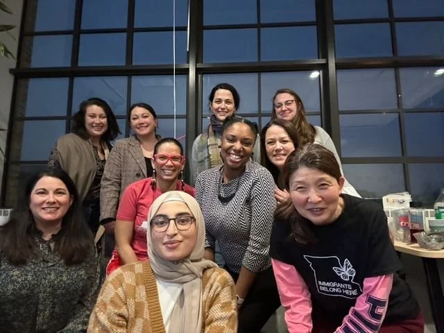 Group of nine women smiling indoors in front of large window, some wearing glasses, diverse in appearance.